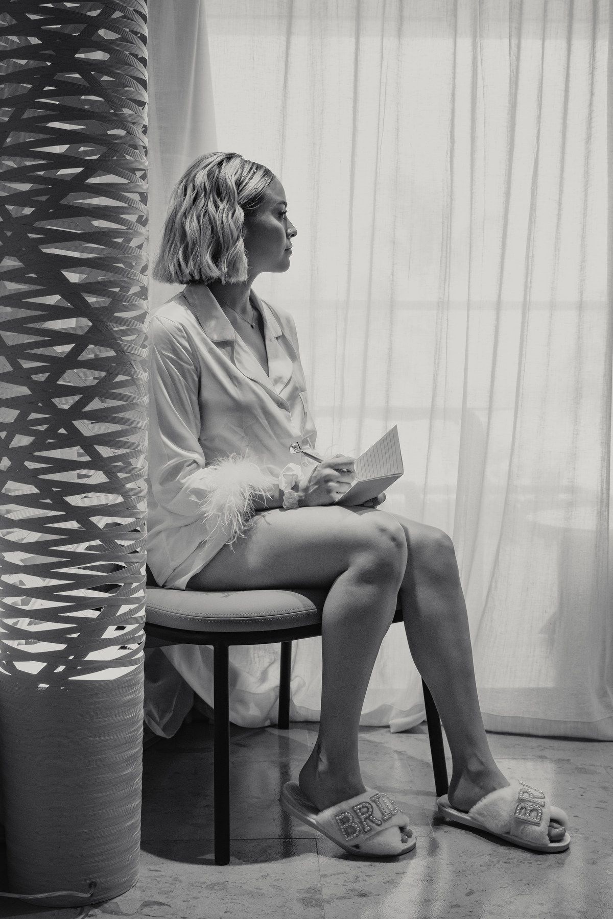 Black and white photo of bride in robe reading vows while seated by sheer curtains.