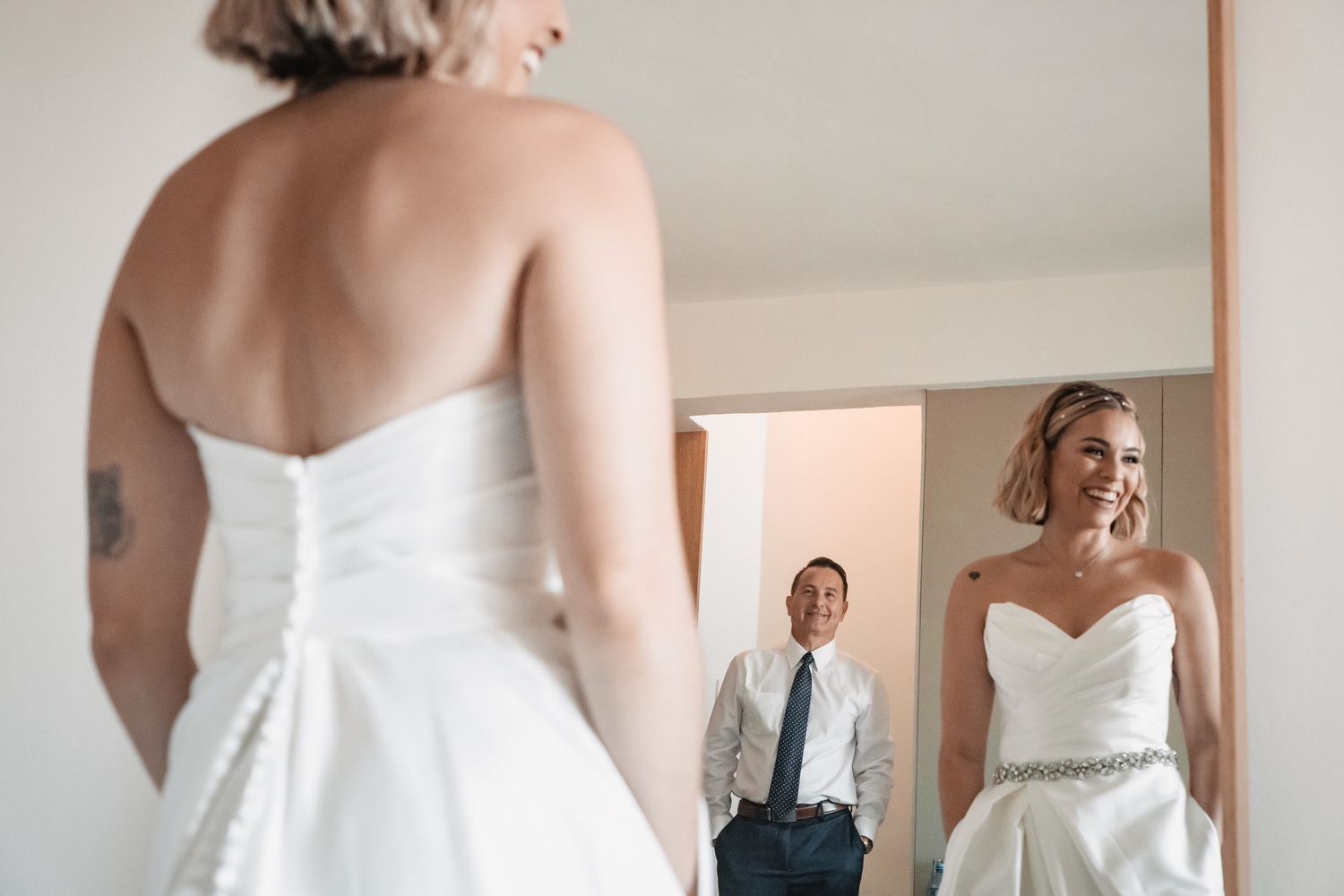 Bride in strapless white gown smiles at her reflection in mirror as groom stands in background during wedding prep.