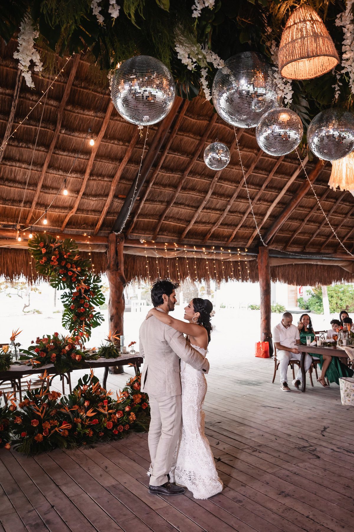 Bride and groom dance under thatched palapa roof adorned with disco balls, string lights, and tropical floral decor.