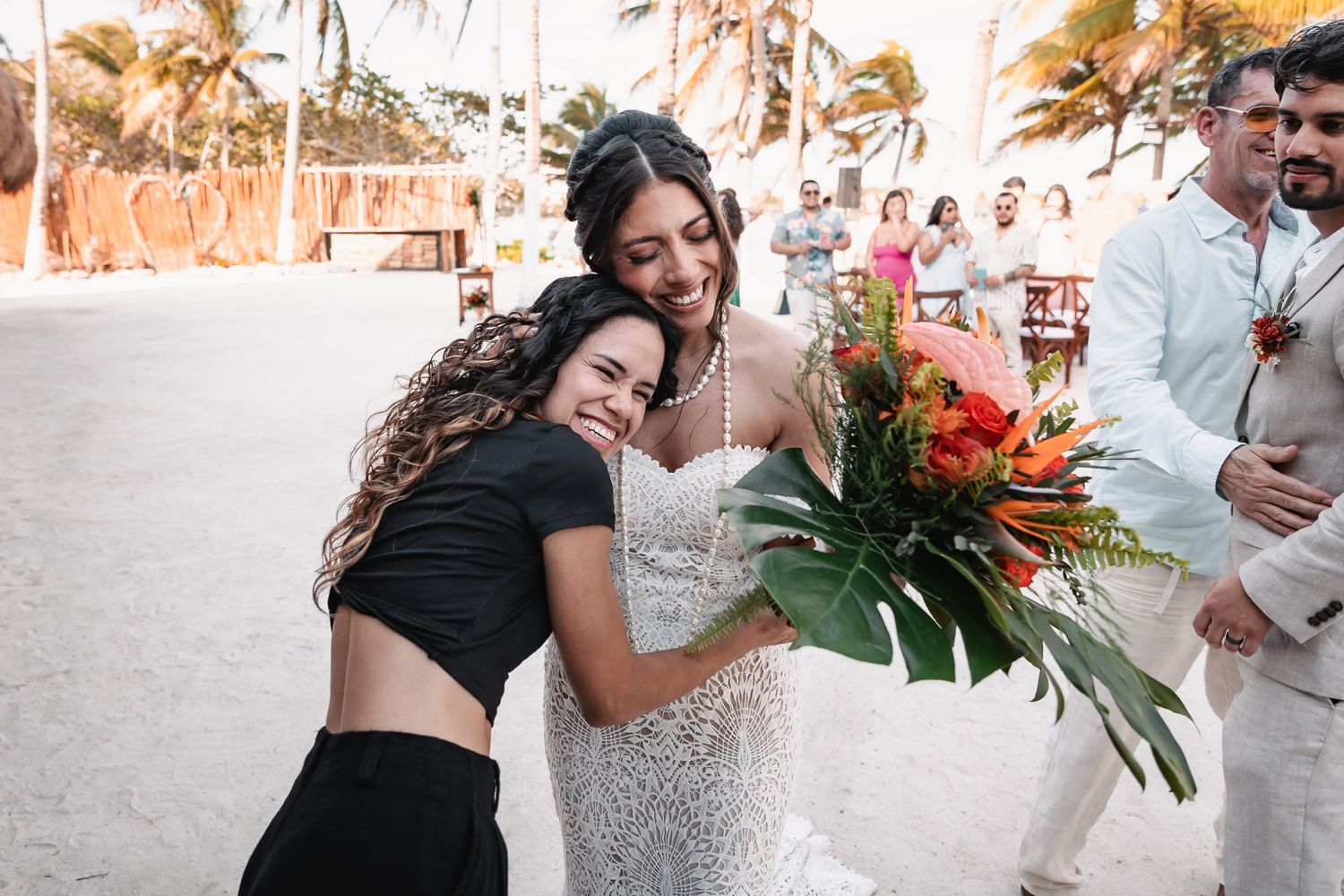 Bride in sparkly gown hugs guest while holding tropical bouquet at beach wedding.