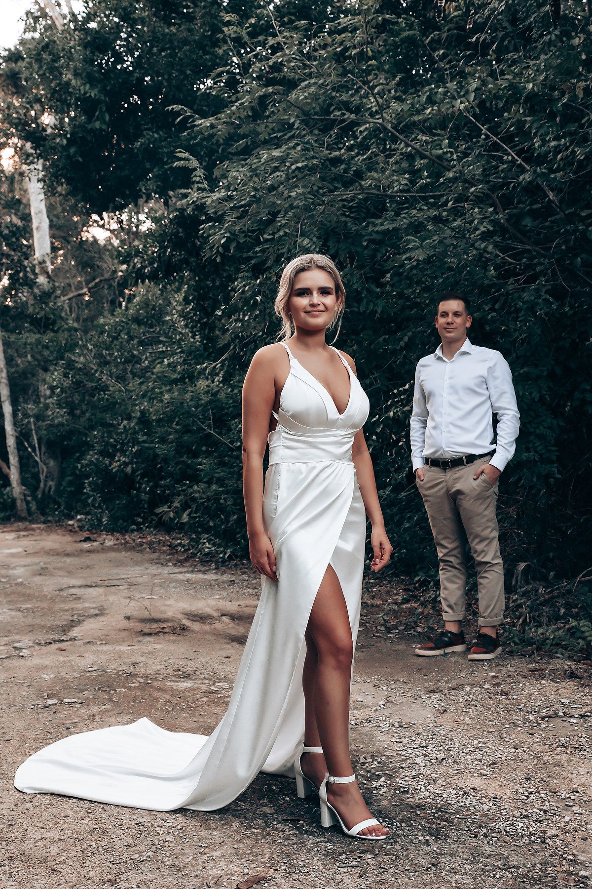 A bride in a flowing white dress poses with her partner on a forest path.