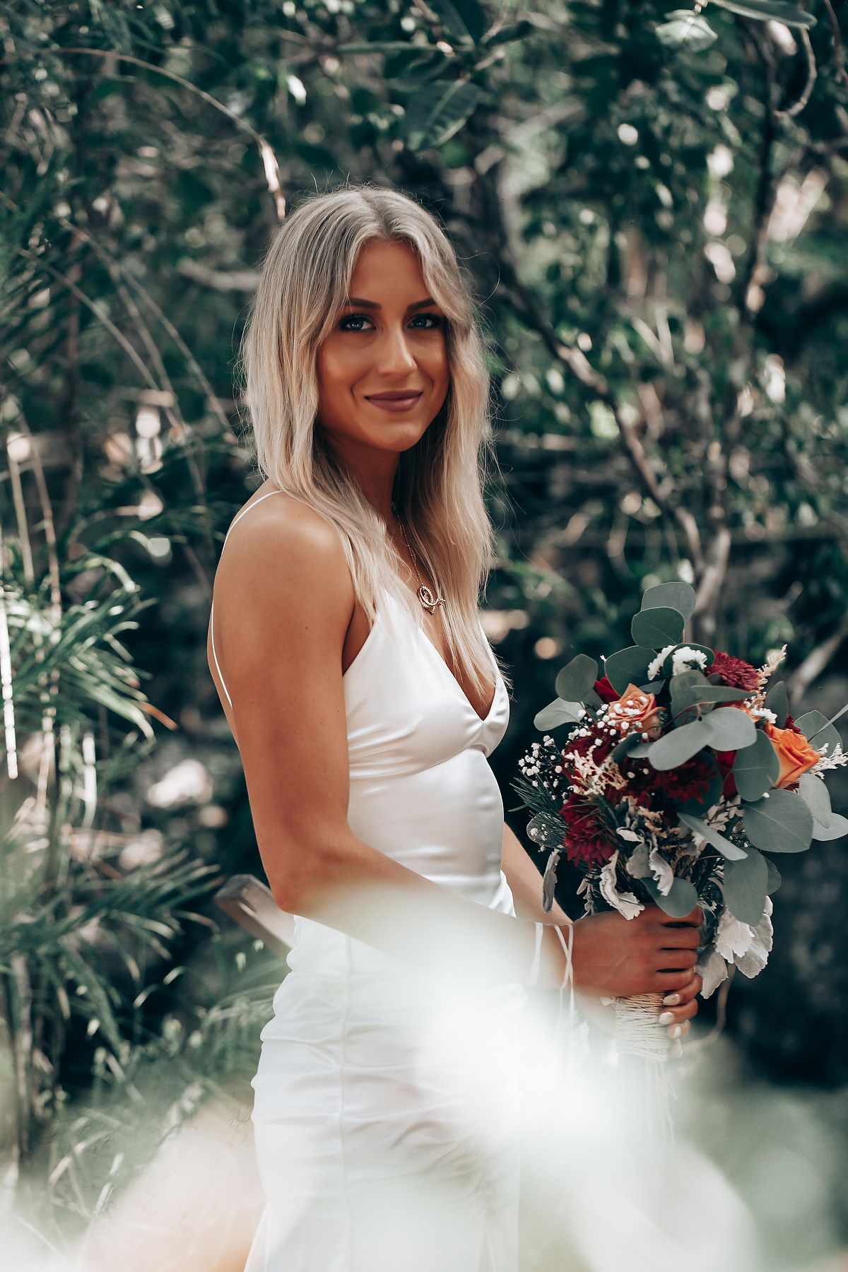A person in a white wedding dress holds a burgundy and white floral bouquet in a forest setting.