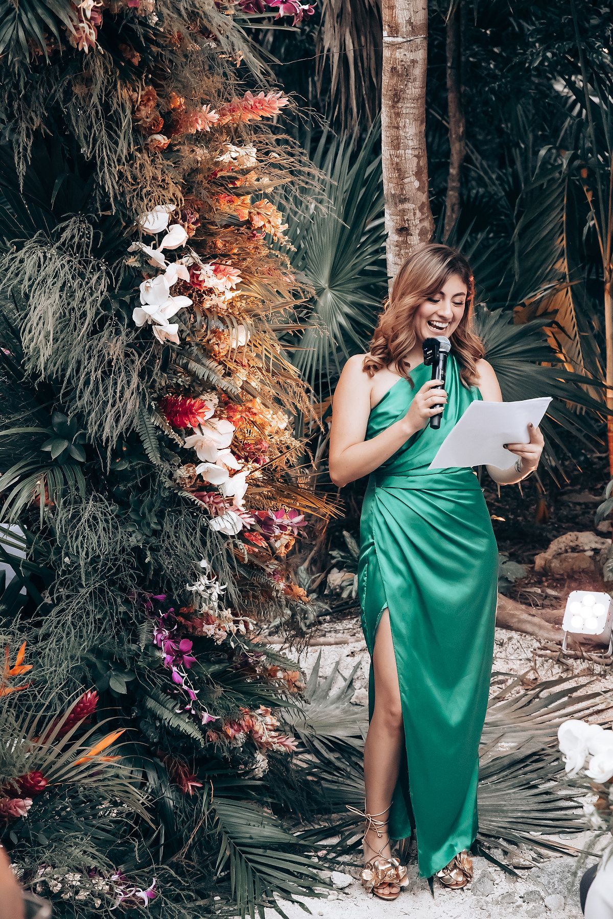 A person in an emerald green dress reads from papers beside a floral arch in a tropical setting.