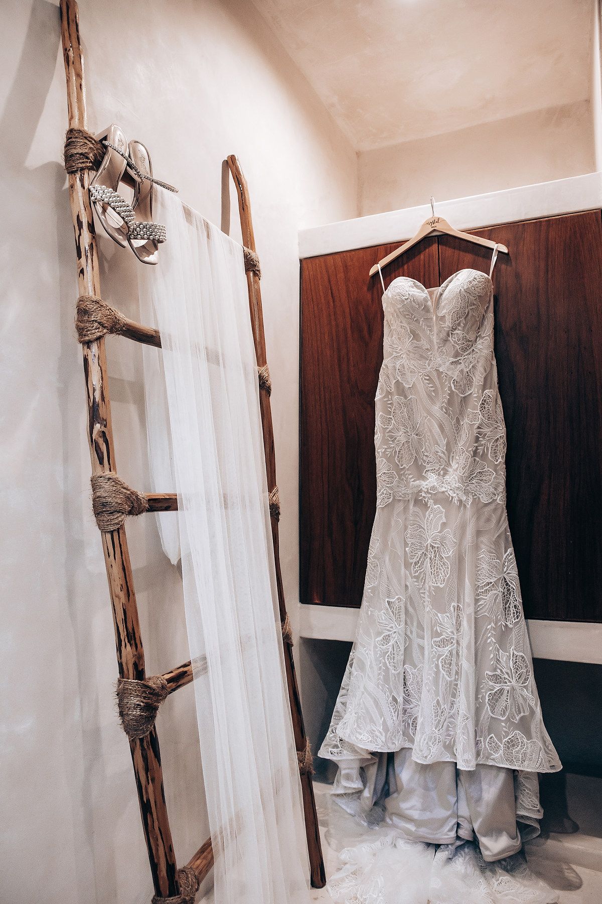 A white lace wedding dress hangs elegantly on a rustic wooden ladder against a cream-colored wall.