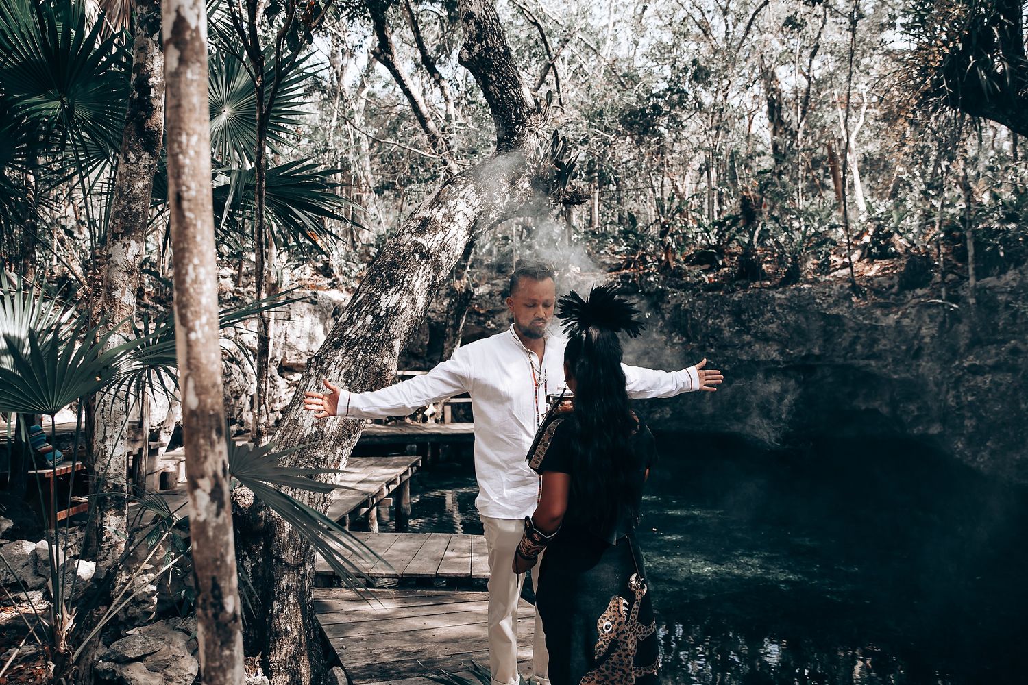 A couple in white outfits poses together in a moody forest setting with dramatic lighting.