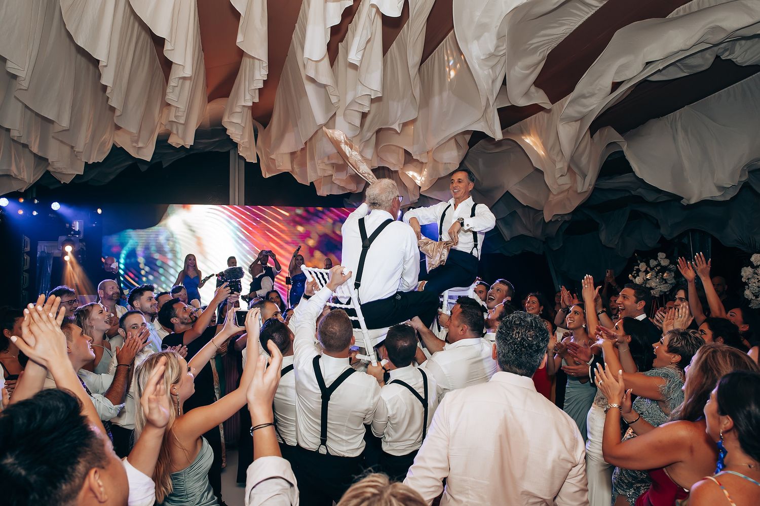 An energetic wedding reception crowd dancing under draped fabric decor.