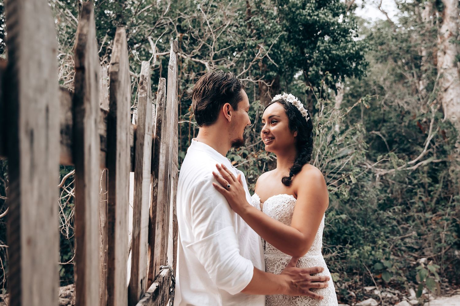 A happy couple in white wedding attire embraces and laughs together in a romantic outdoor setting with tall trees.