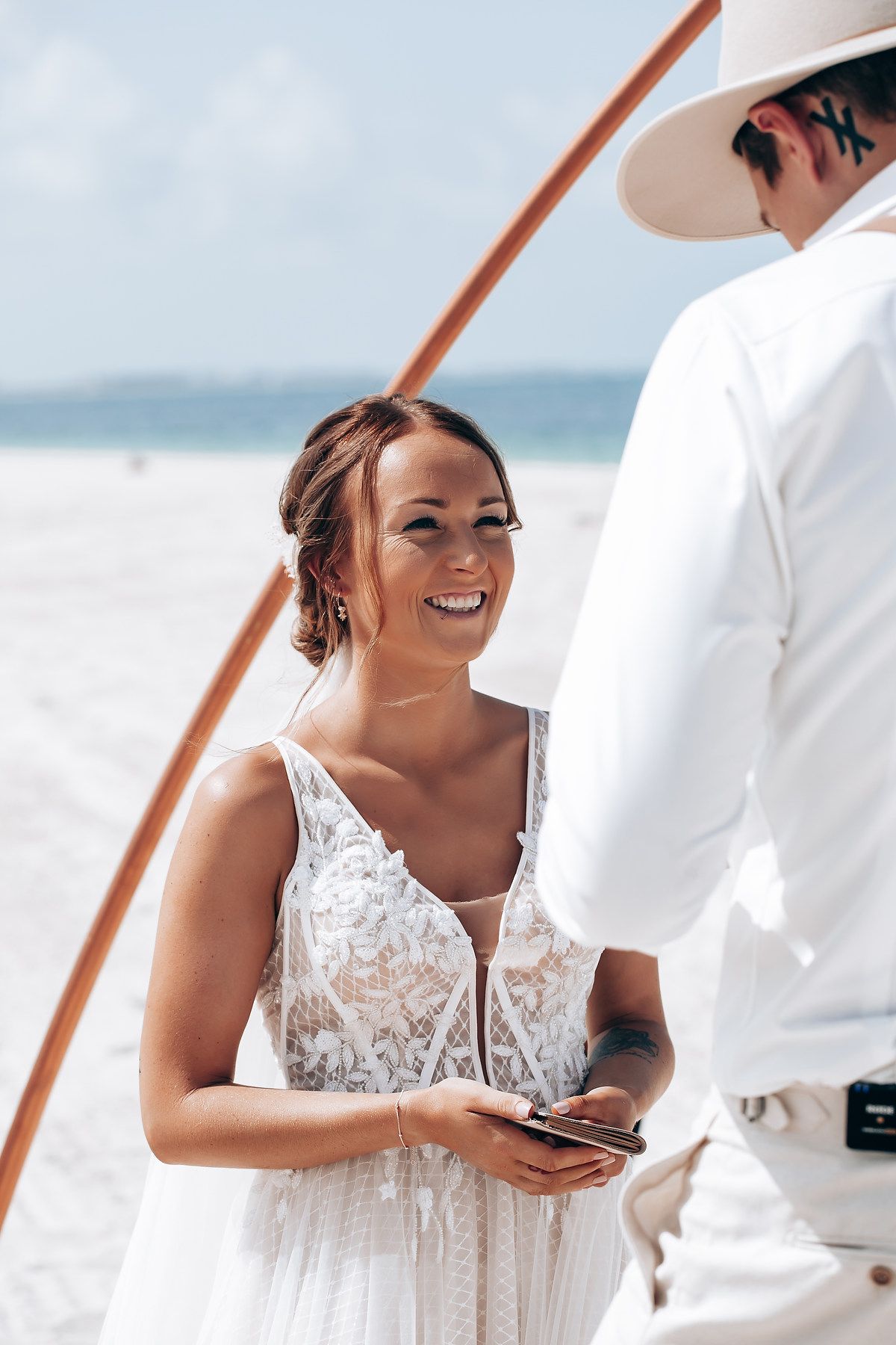 Bride in white lace dress smiling during a beach wedding ceremony.