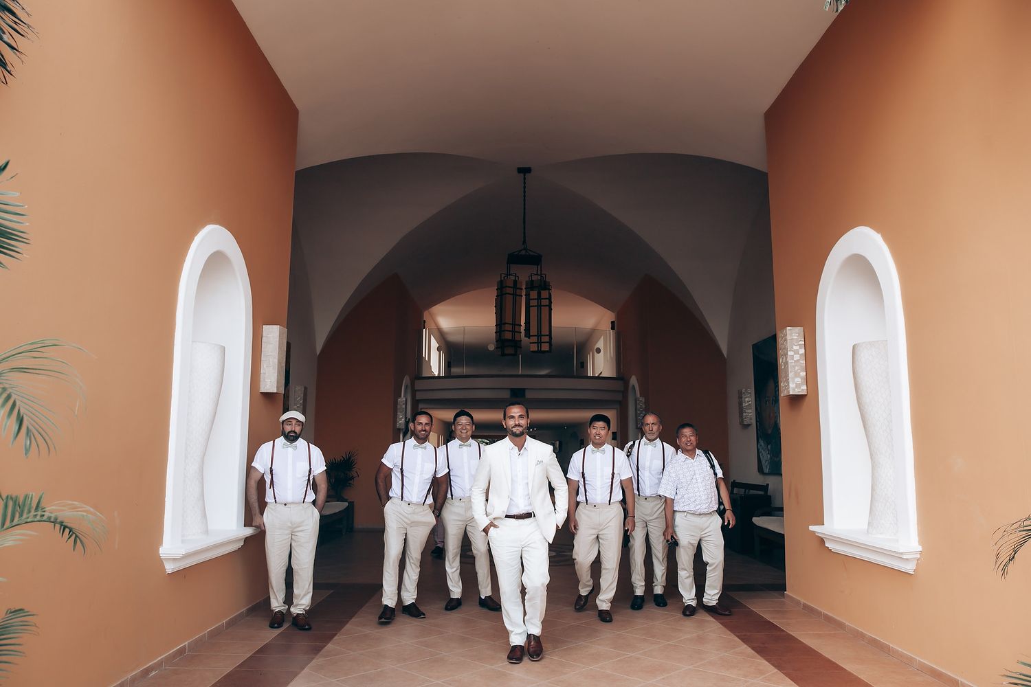 Wedding party members in white outfits walk through an arched hallway.