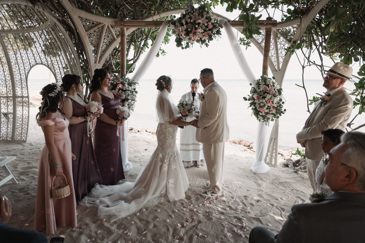 Wedding ceremony under decorative arch on beach with bridesmaids in mauve dresses and floral arrangements.