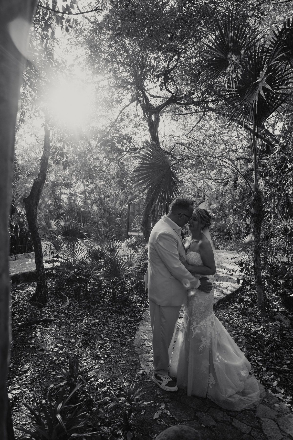 Black and white photo of a bride and groom embracing in a lush garden with sunlight filtering through trees.