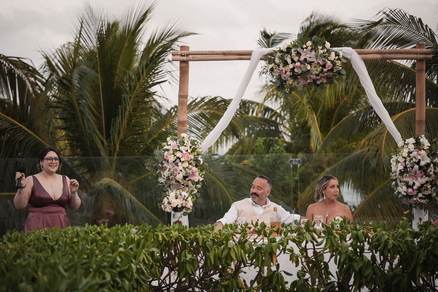 Bride and groom seated at ceremony altar beneath floral arch with palm trees in background.