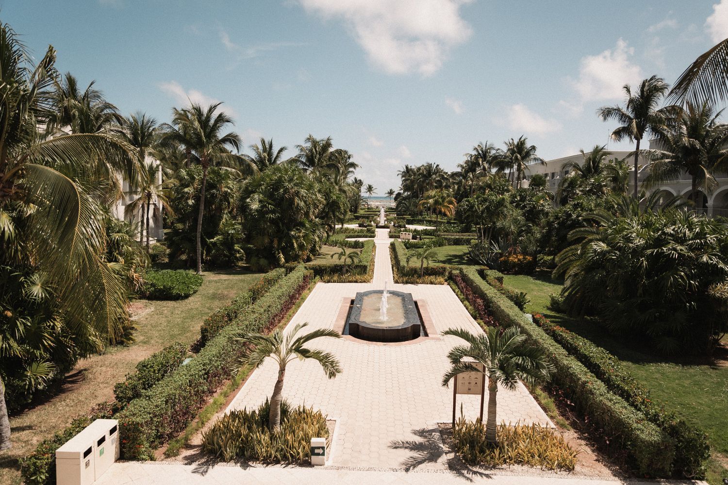 Lush tropical garden pathway with fountain, palm trees, and manicured hedges at a luxury resort.