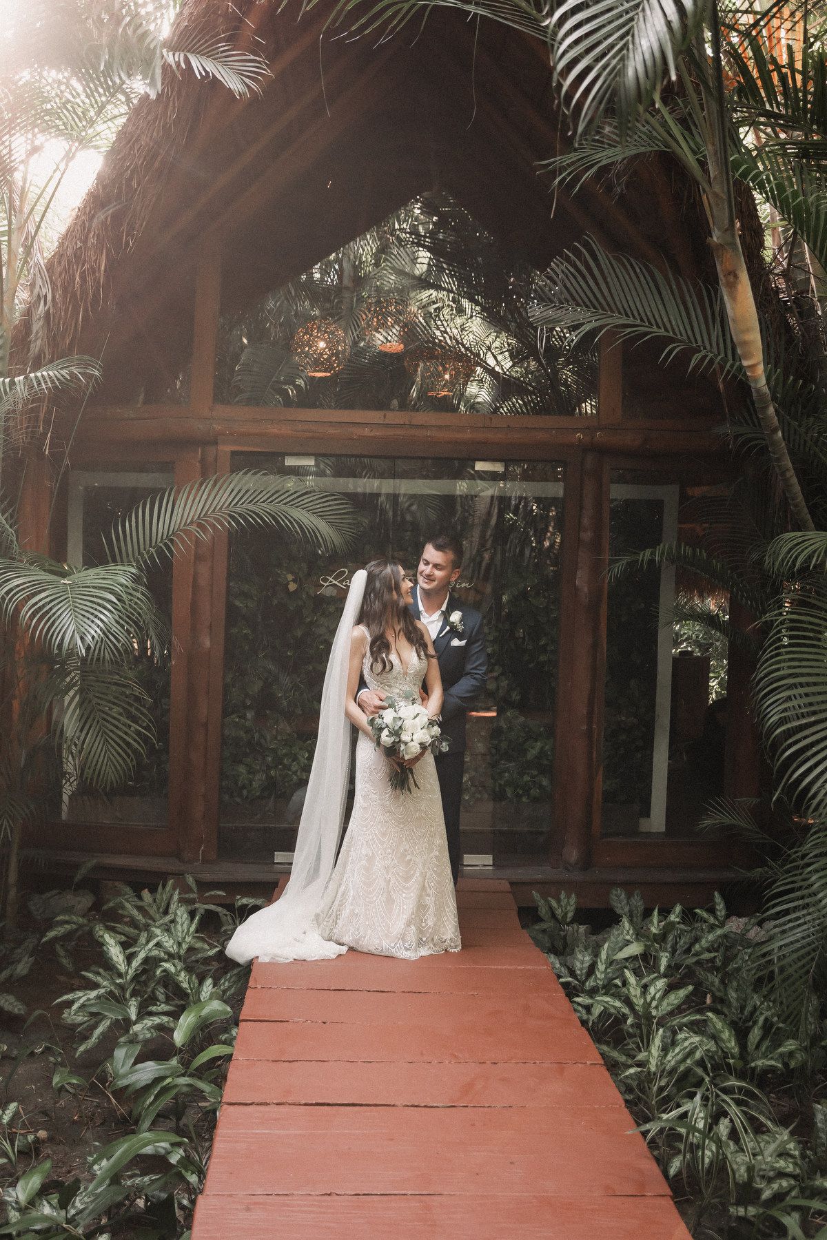 Bride and groom pose on red wooden path surrounded by lush tropical greenery and palms.