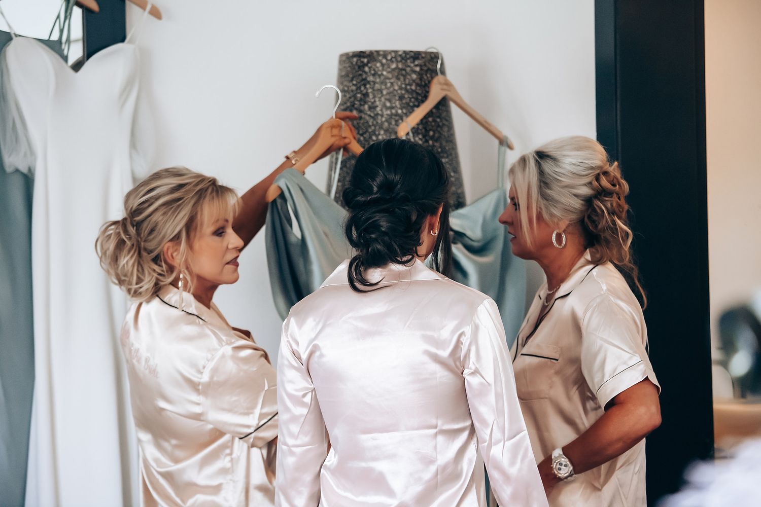 Bridal party in matching white satin robes getting ready together in a bright dressing room.