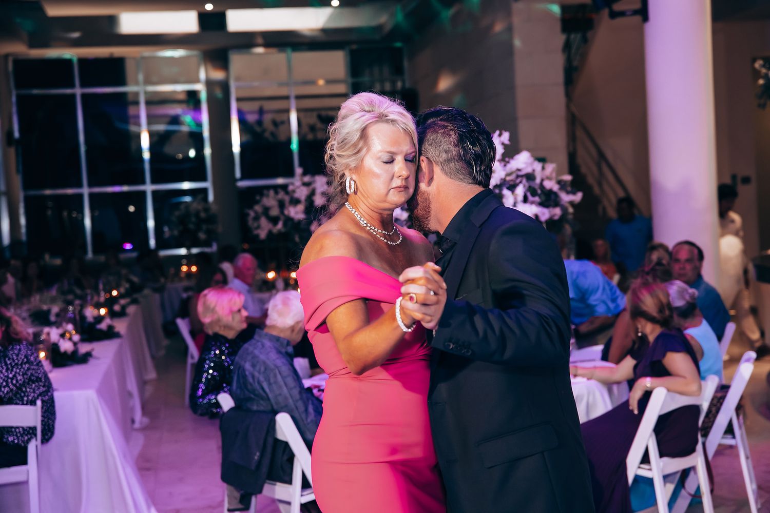 A couple dances together in formal attire under pink and purple lighting at an evening reception.