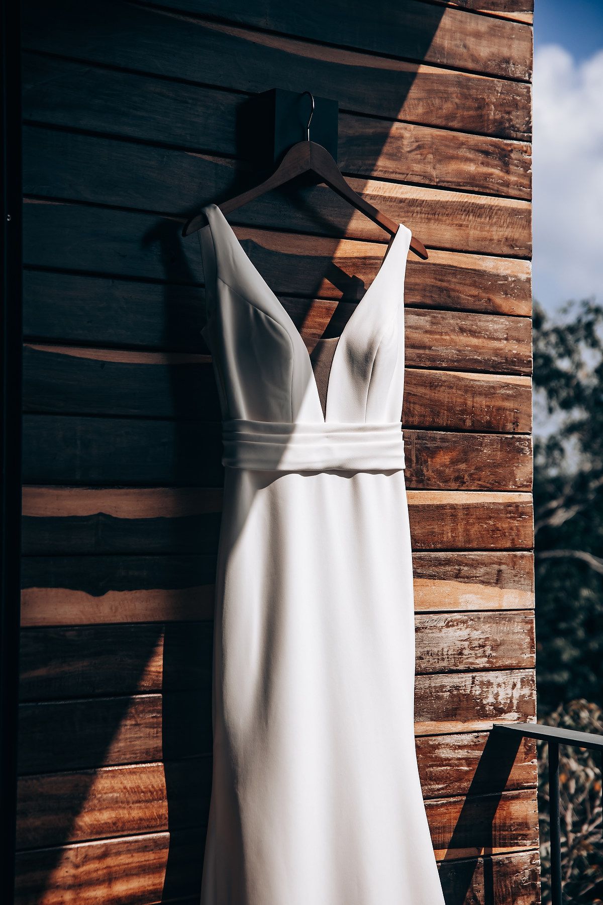 A white wedding dress hangs elegantly against a rustic wooden wall backdrop in dramatic lighting.