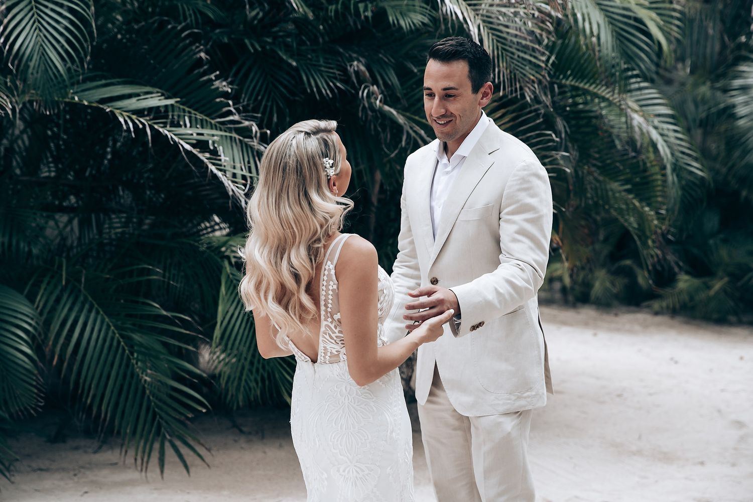 A couple in white attire share a romantic moment among tropical palm trees.