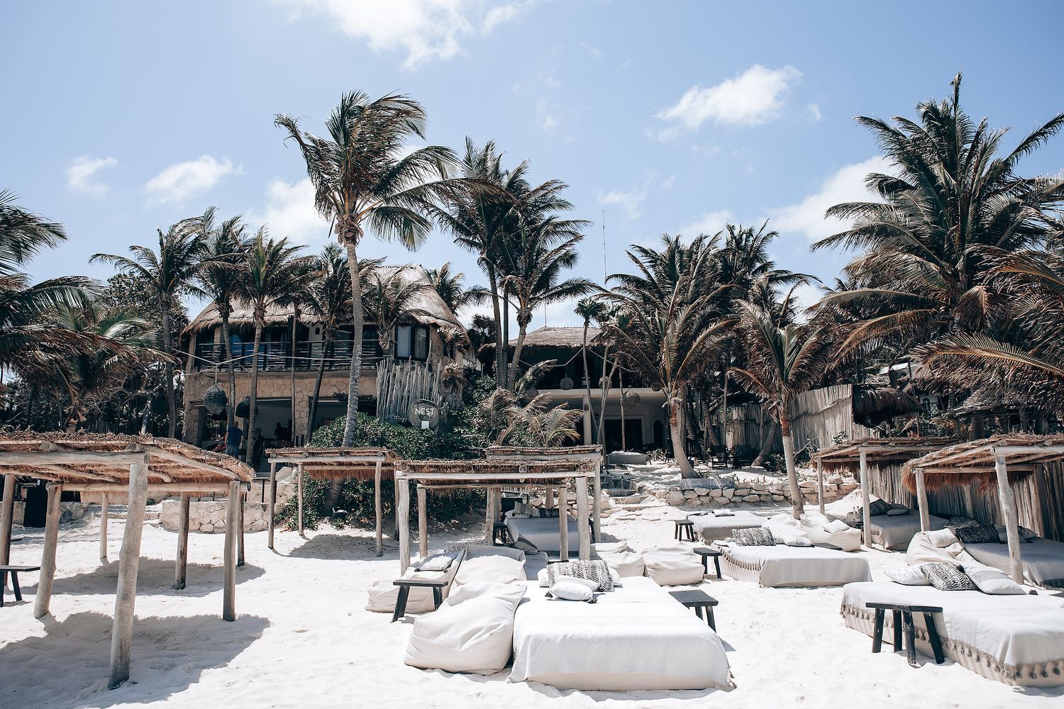 Beachfront resort with white sand, palm trees, and thatched-roof cabanas along a tropical coastline.