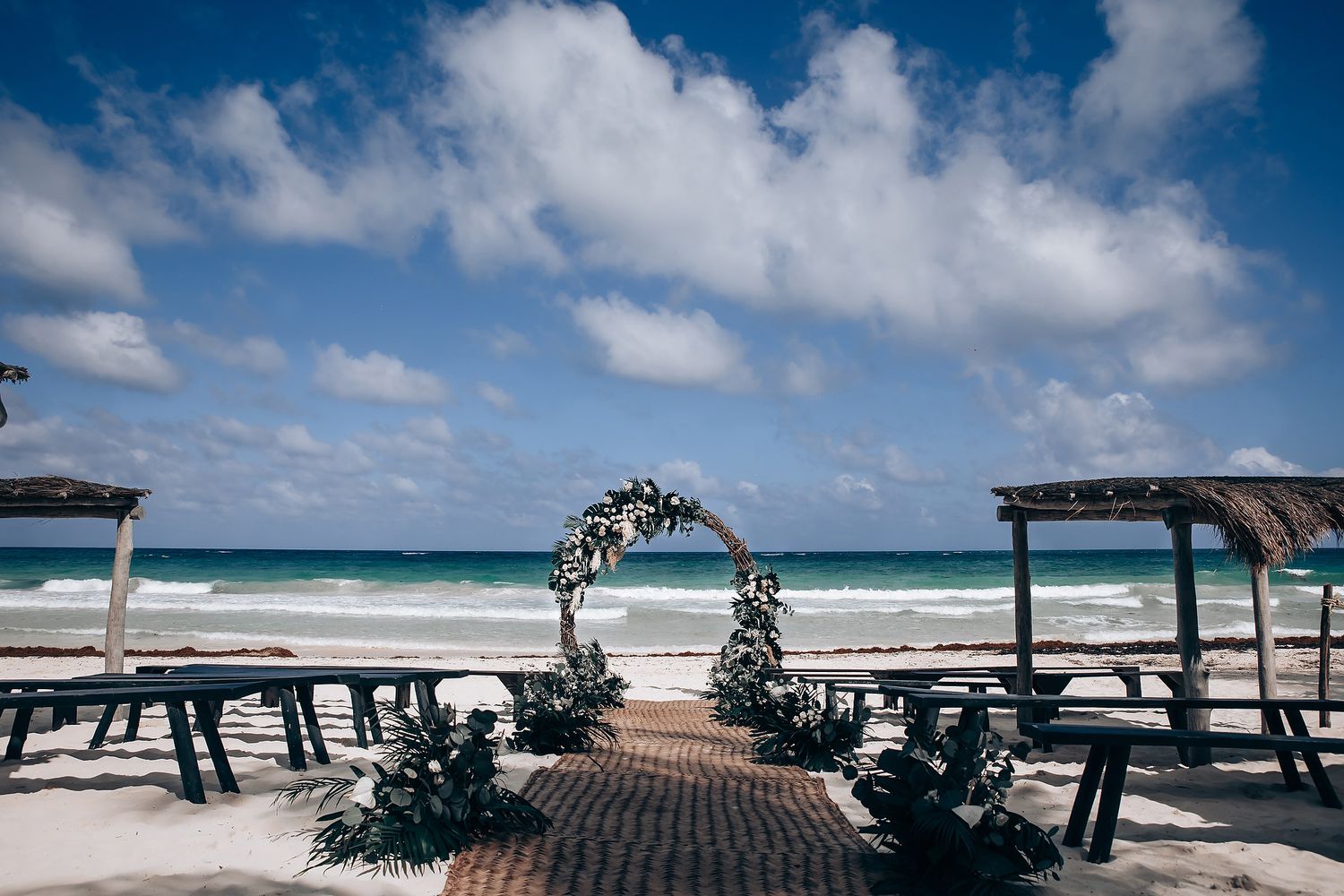 Beachfront wedding ceremony setup with white floral arch against blue sky and ocean waves.