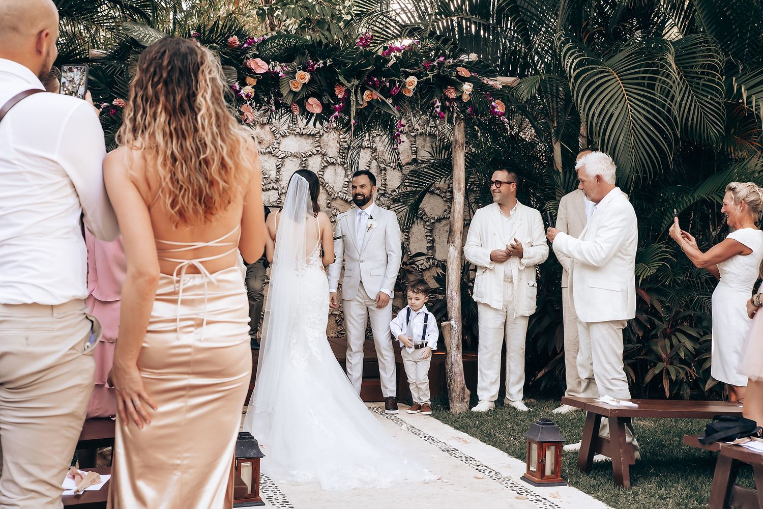 Wedding guests in light colored attire gather outdoors for a celebration under palm trees.