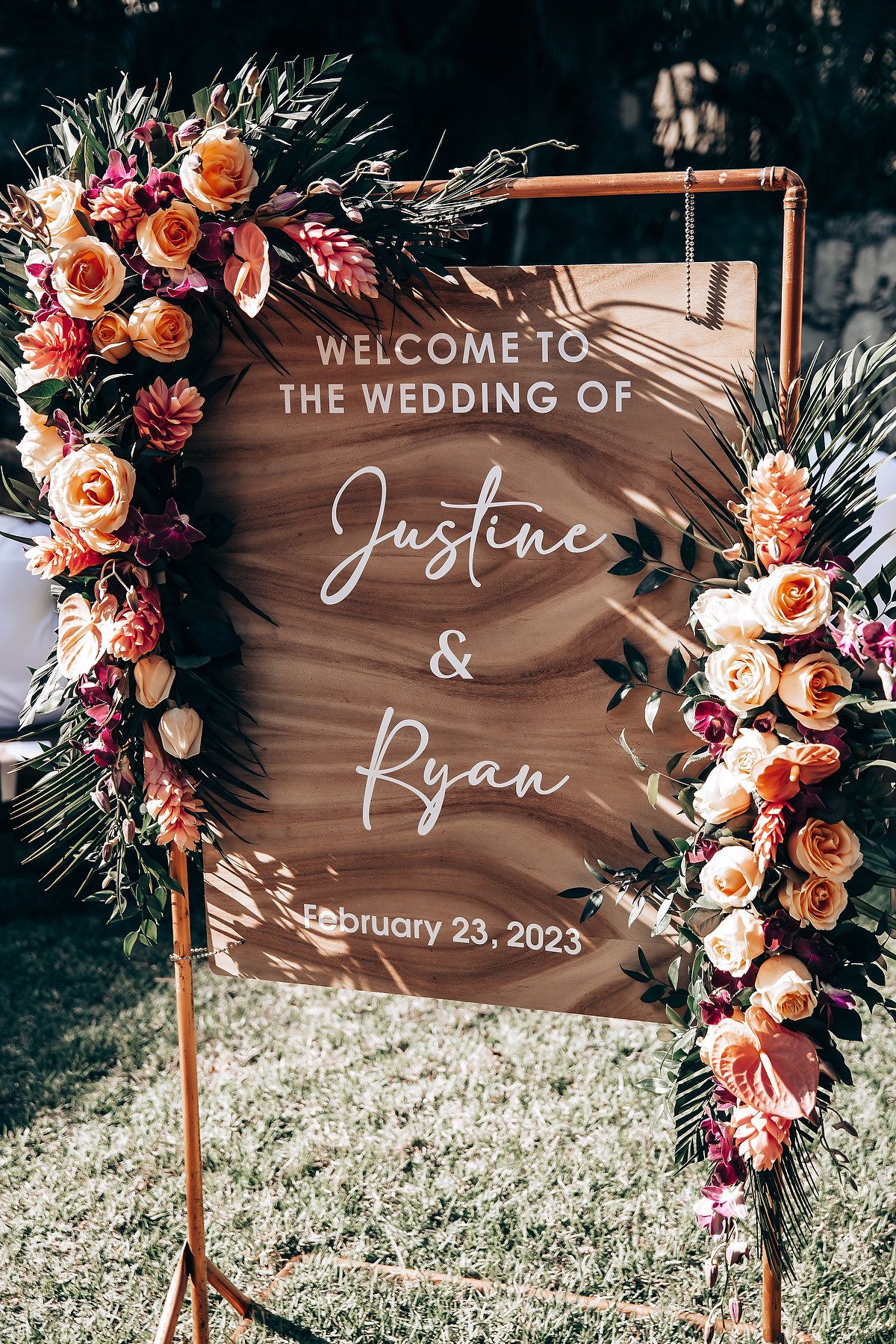 Wooden welcome sign decorated with coral and peach flowers at an outdoor wedding venue.