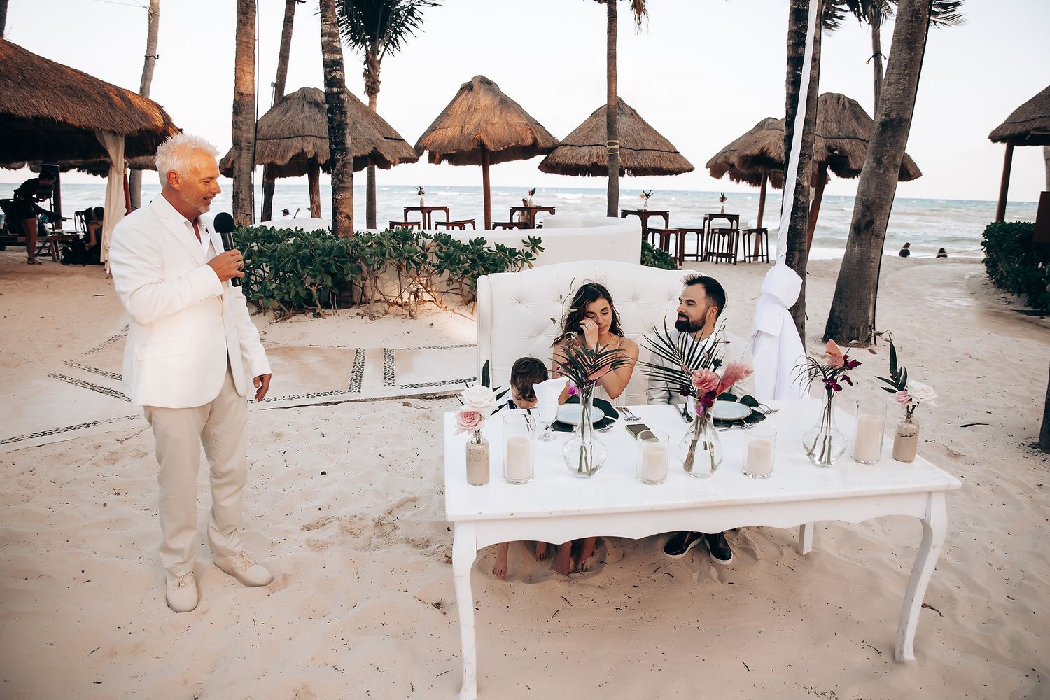 Beach wedding reception setup with thatched umbrellas and white furniture on sand.