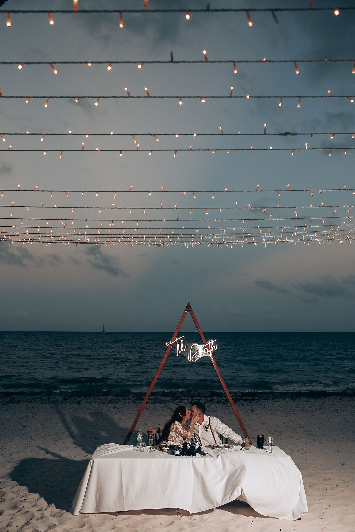 Beach wedding reception under string lights with white tablecloths and ocean backdrop at dusk.