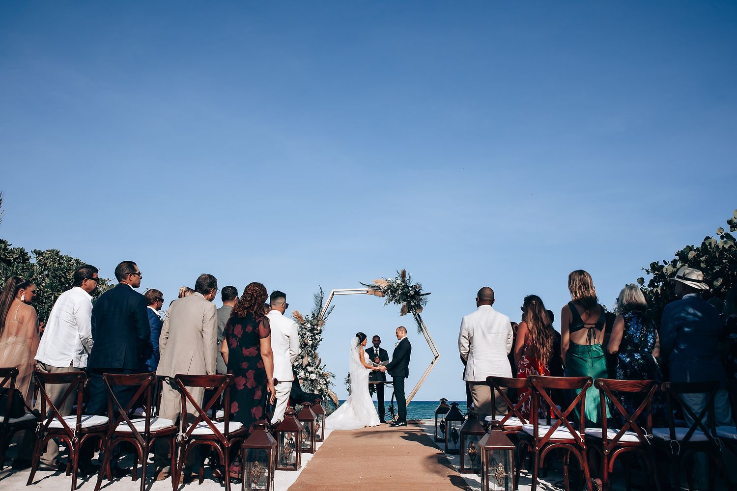 Wedding guests seated outdoors under blue skies watching a beachside ceremony.