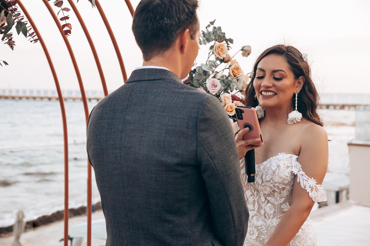 Romantic wedding ceremony by the ocean with floral arch and sunset lighting.