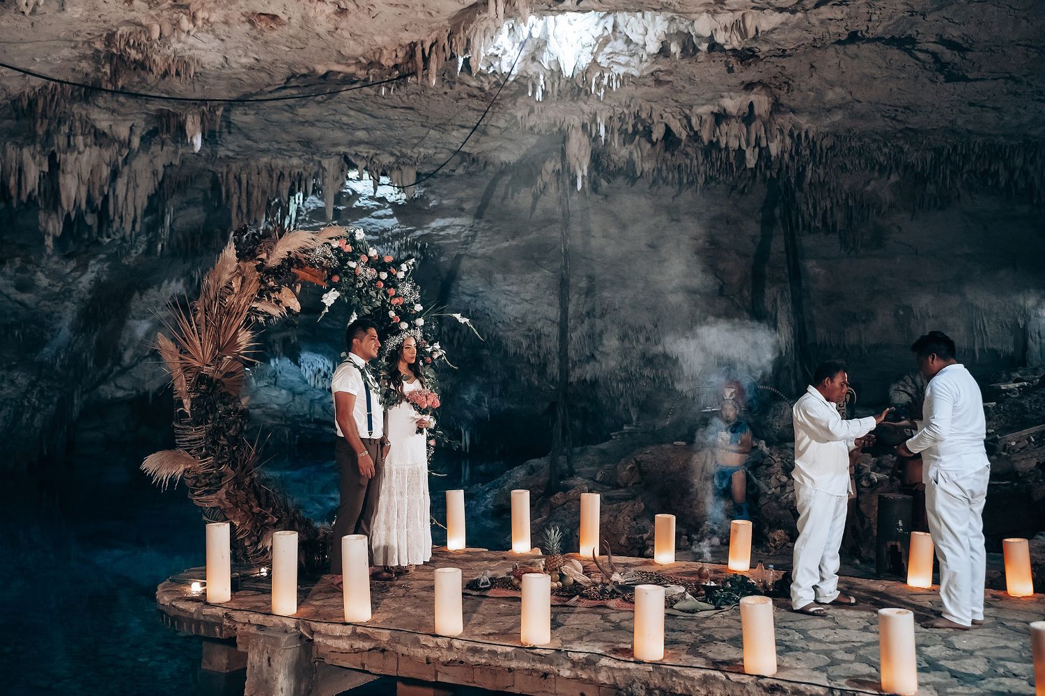 People in white robes walk through an ancient cave system with illuminated stone columns and dramatic lighting.