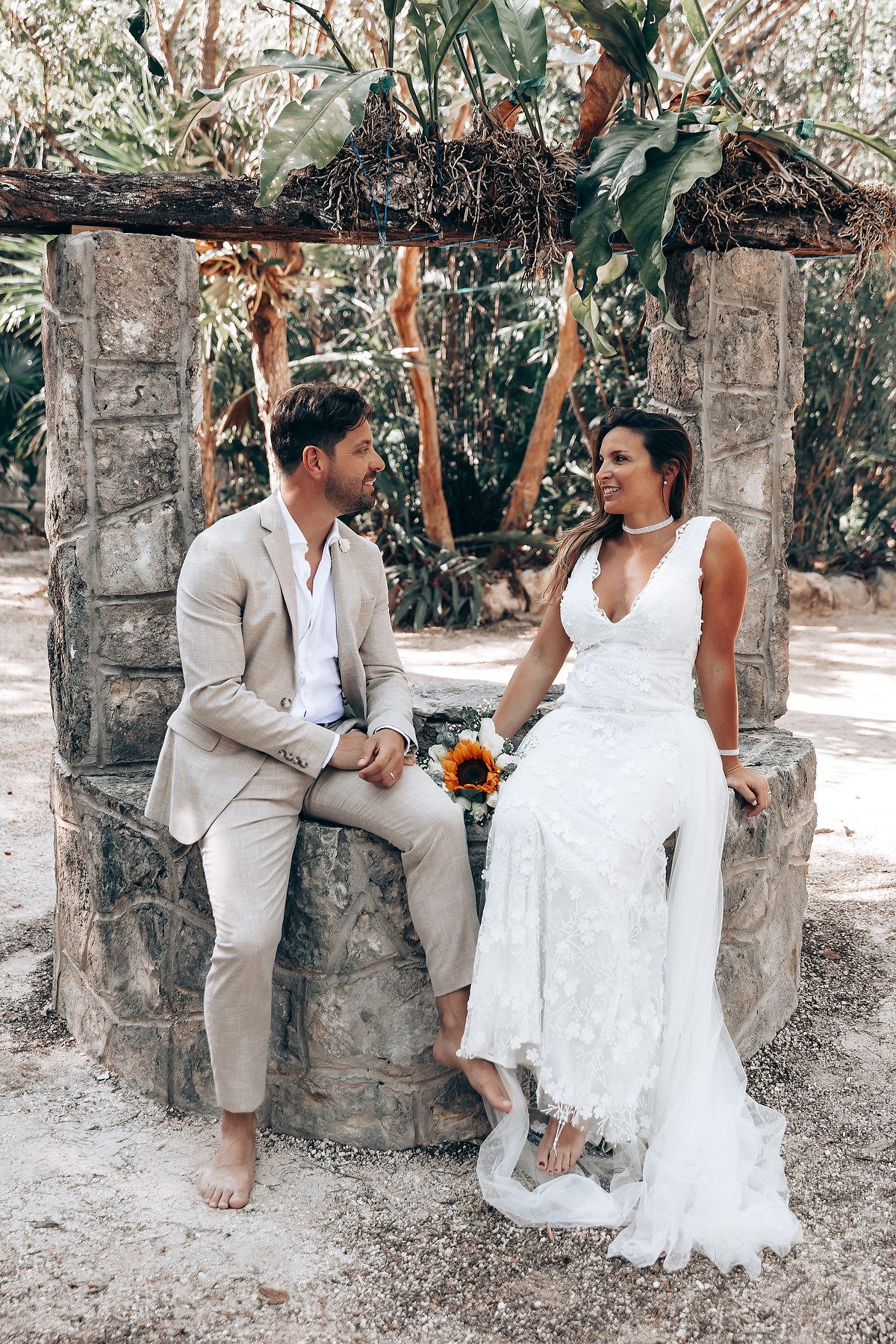 A playful wedding photoshoot with the couple posing on a rustic stone wall surrounded by greenery.