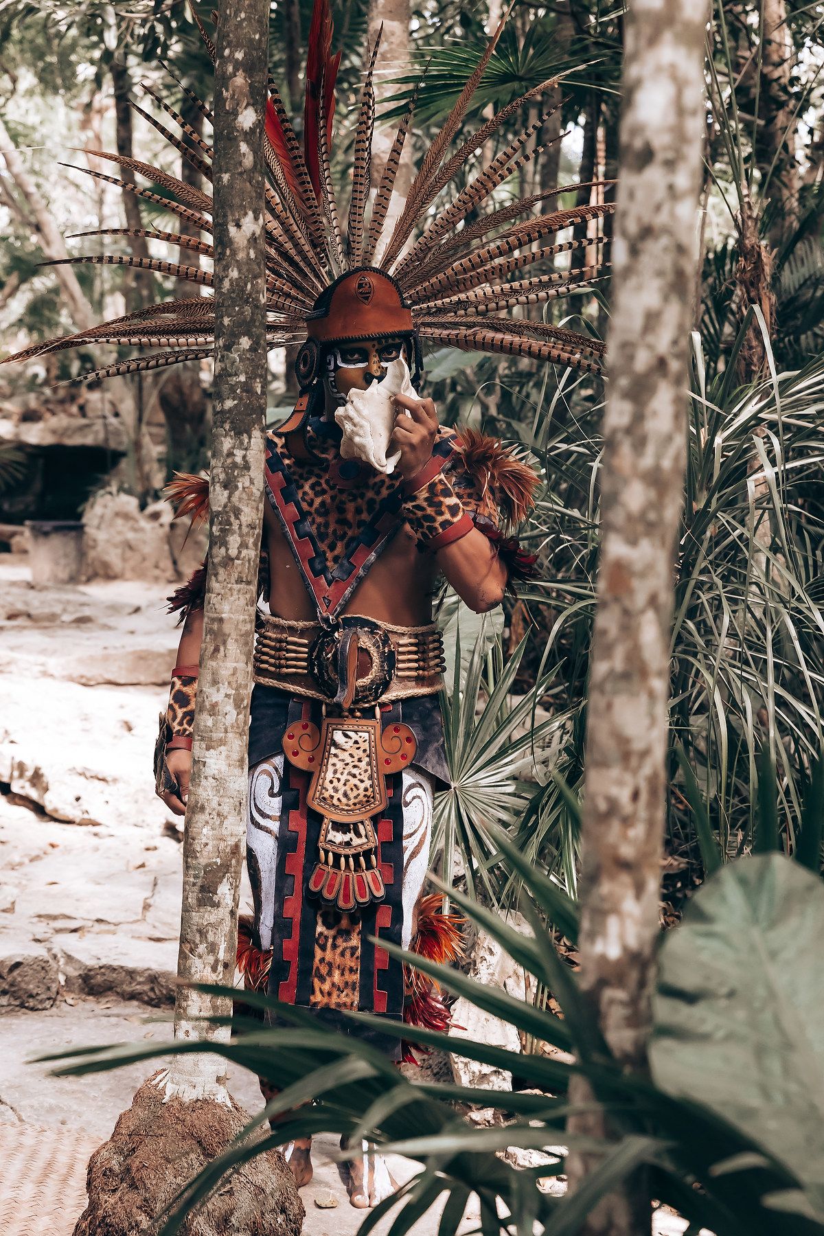 A person wearing traditional Mayan ceremonial attire and feathered headdress poses in a forest setting.