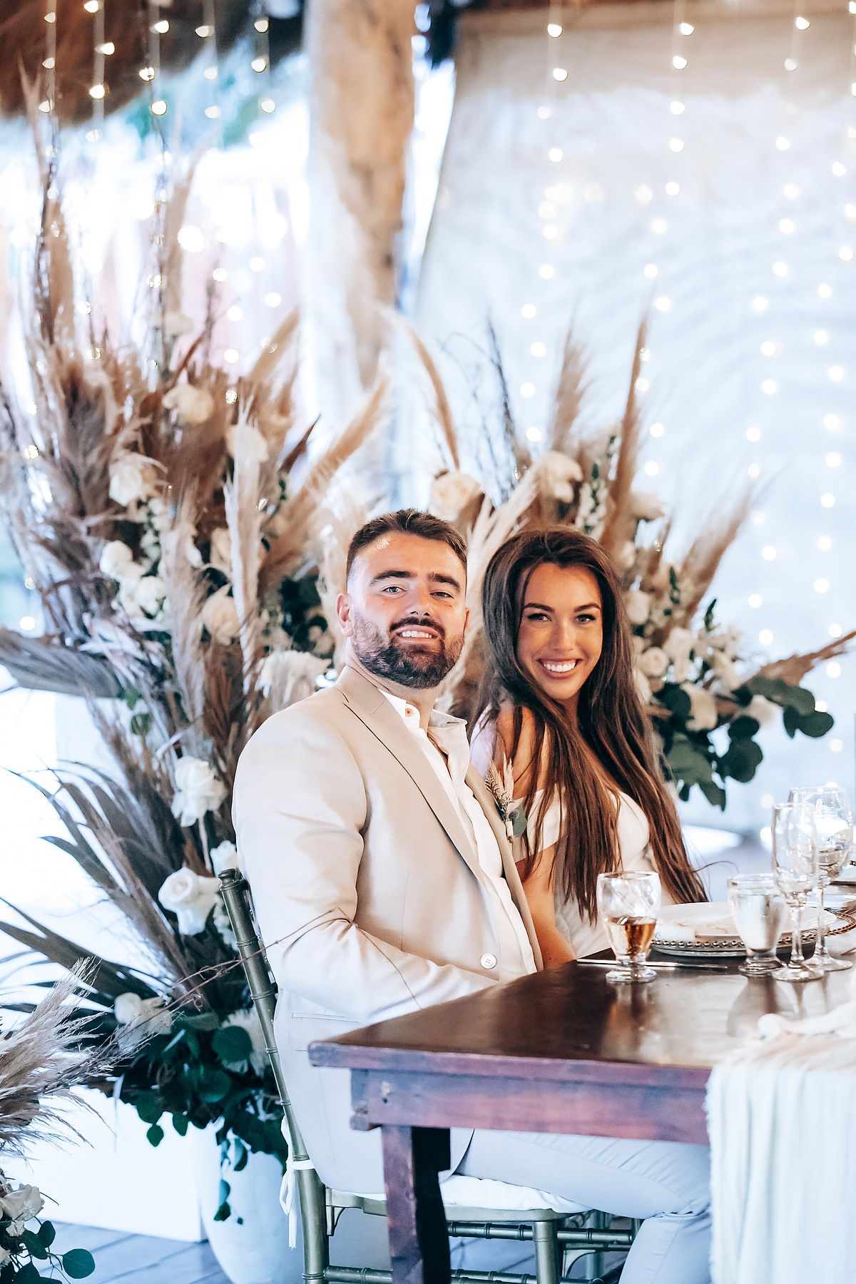 Couple sitting at elegant table with dried floral arrangements and sparkling lights in romantic restaurant setting.