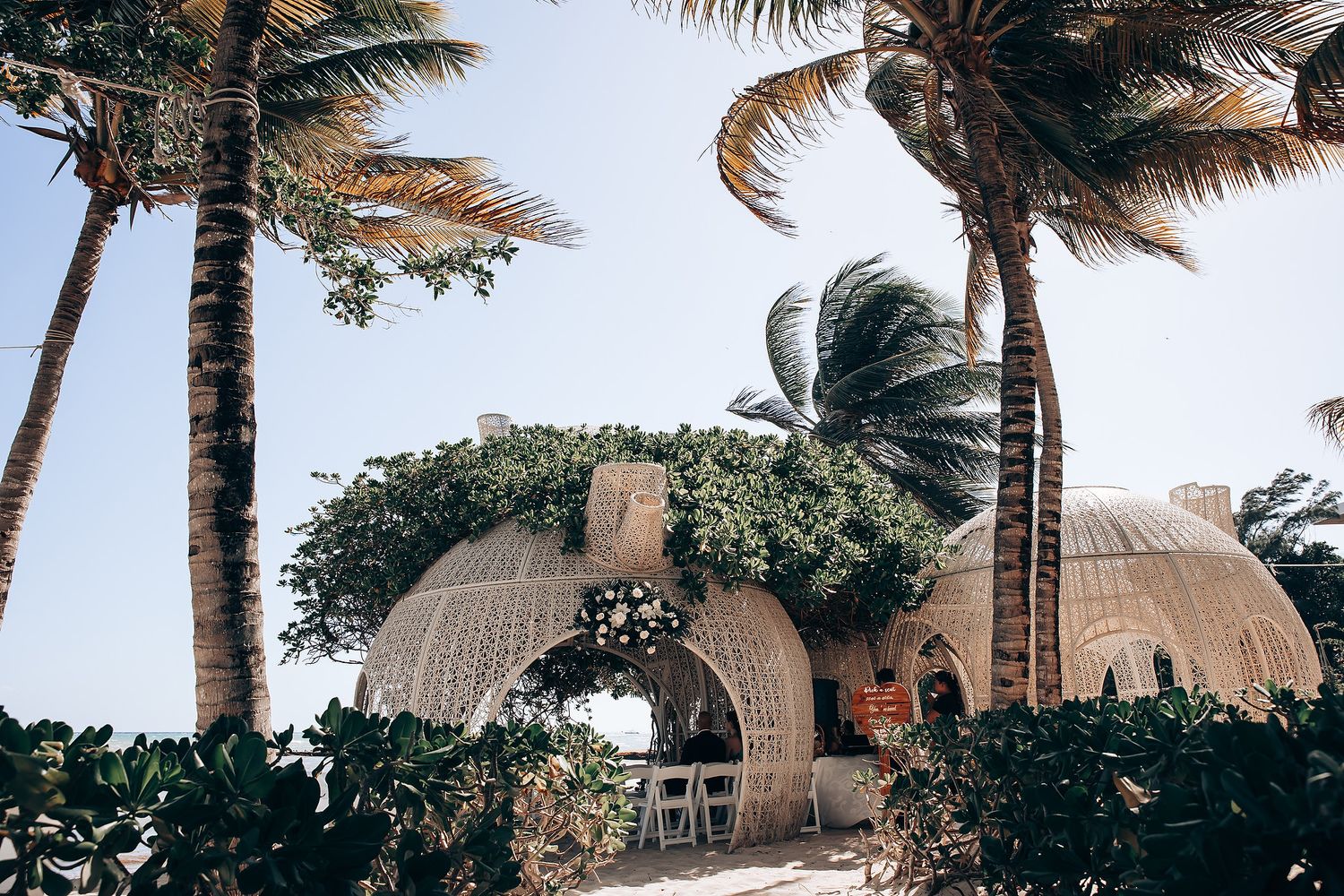 Palm trees sway in the wind at a tropical beach wedding venue with thatched roof structures.