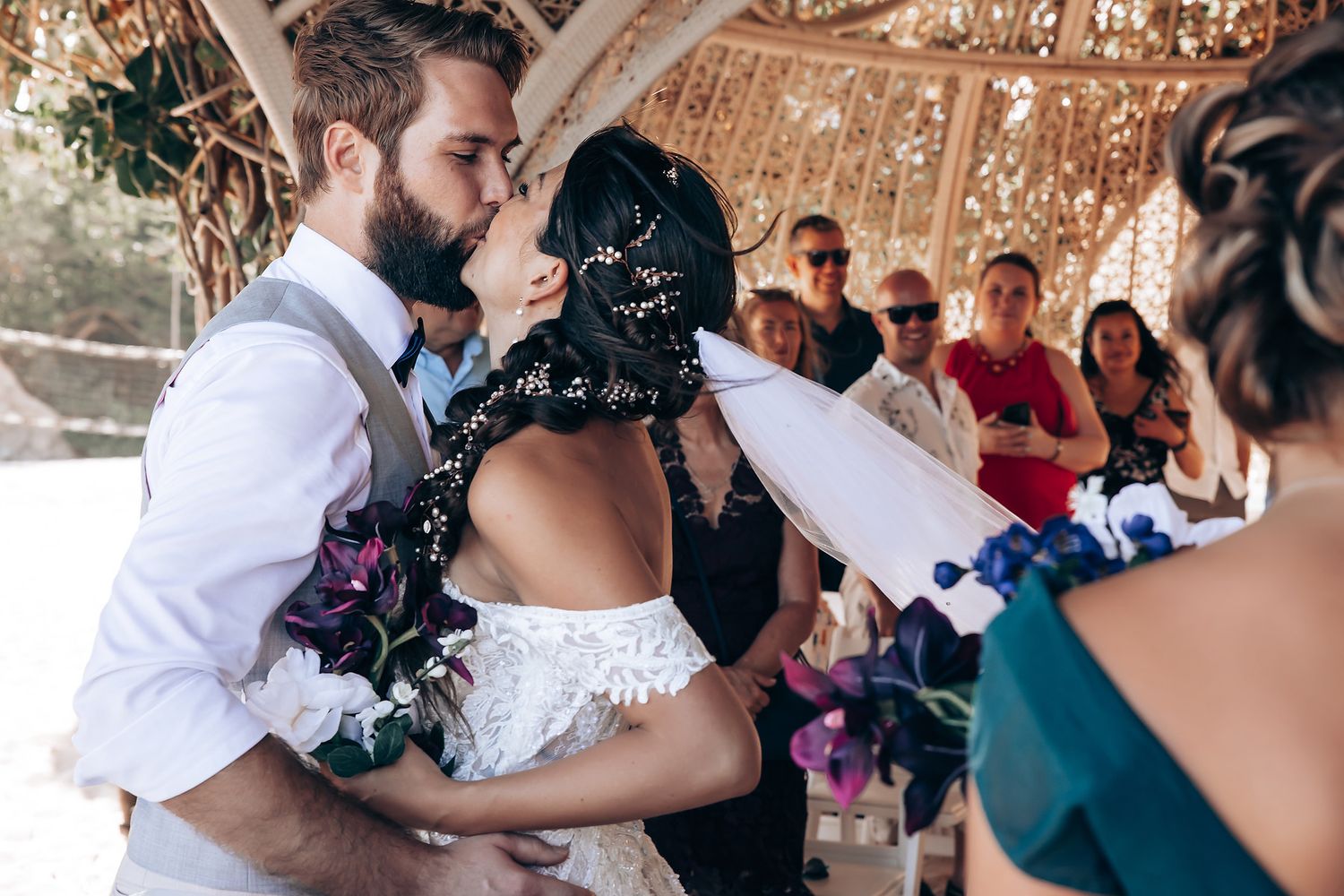Wedding couple shares their first kiss under a decorative pergola surrounded by guests.