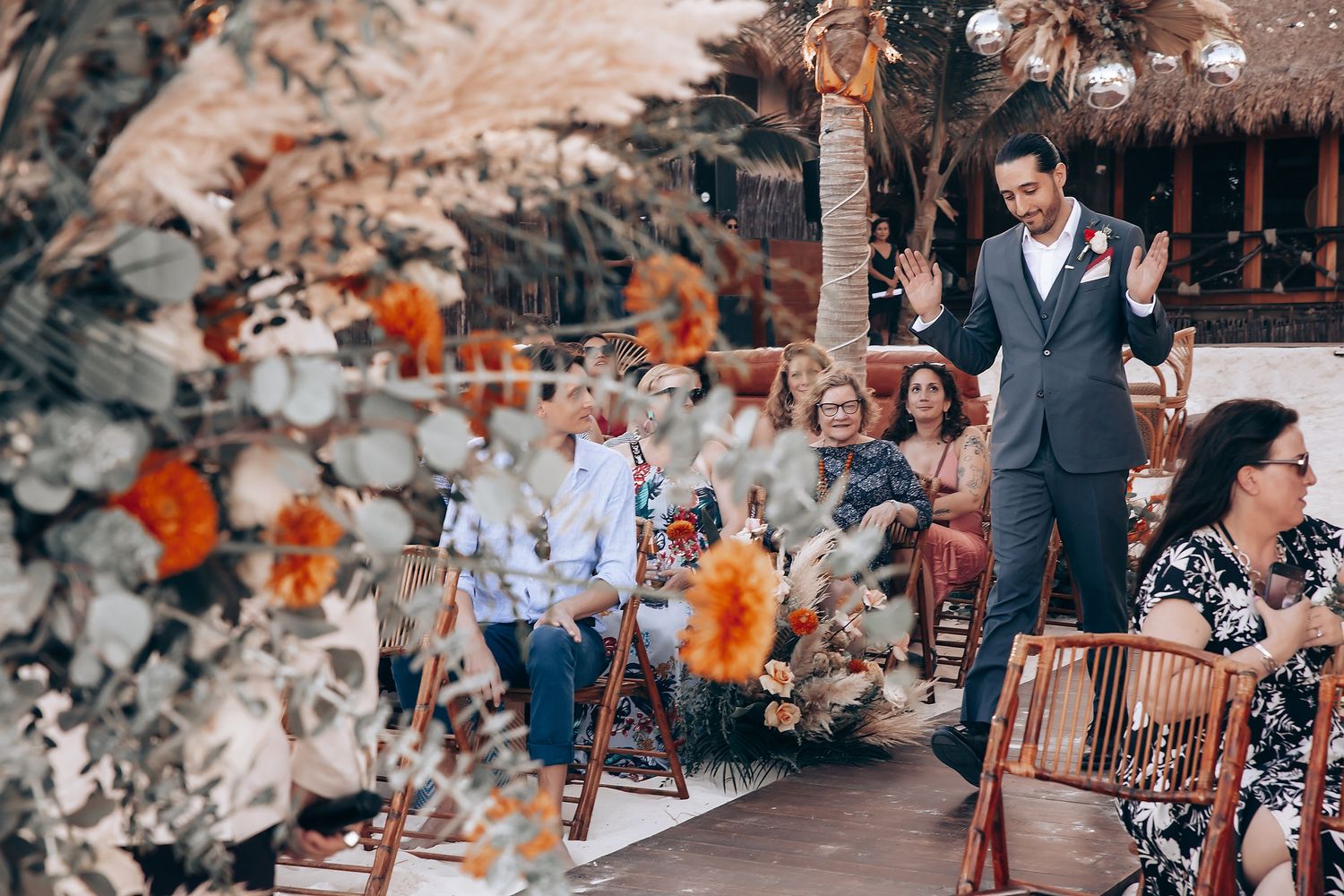 Wedding guests seated at tables decorated with orange florals under palm trees at a beachside venue.