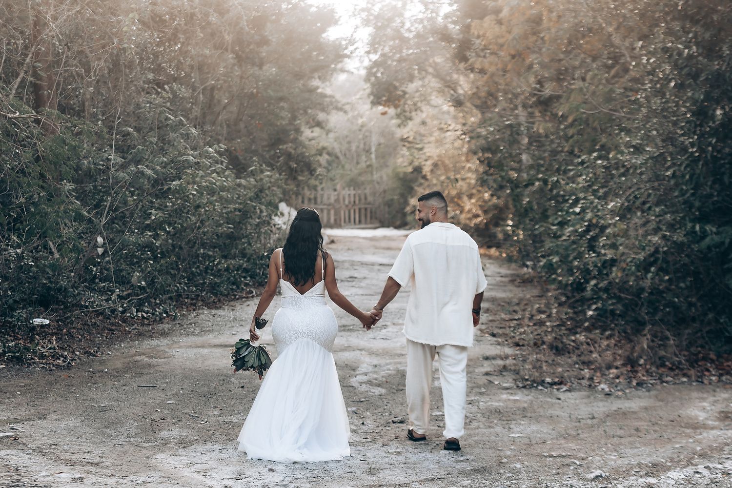 A couple walks hand in hand down a forest path in matching white wedding attire.