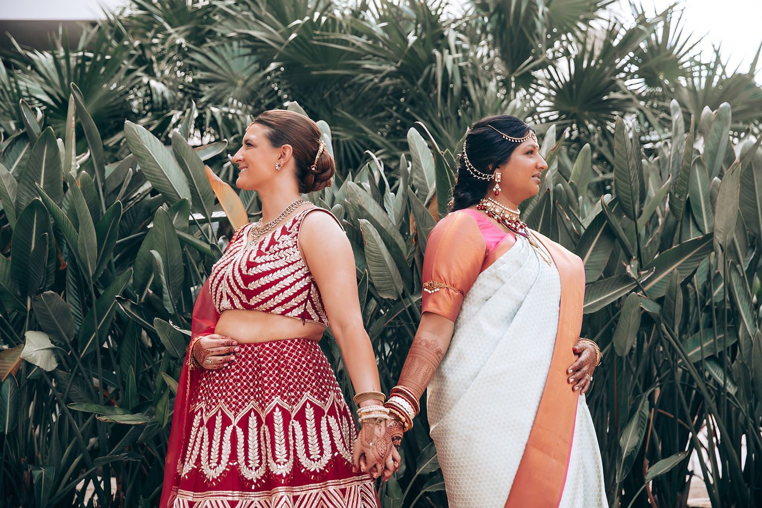 Two people in contrasting traditional and modern outfits pose back-to-back in a tropical setting.