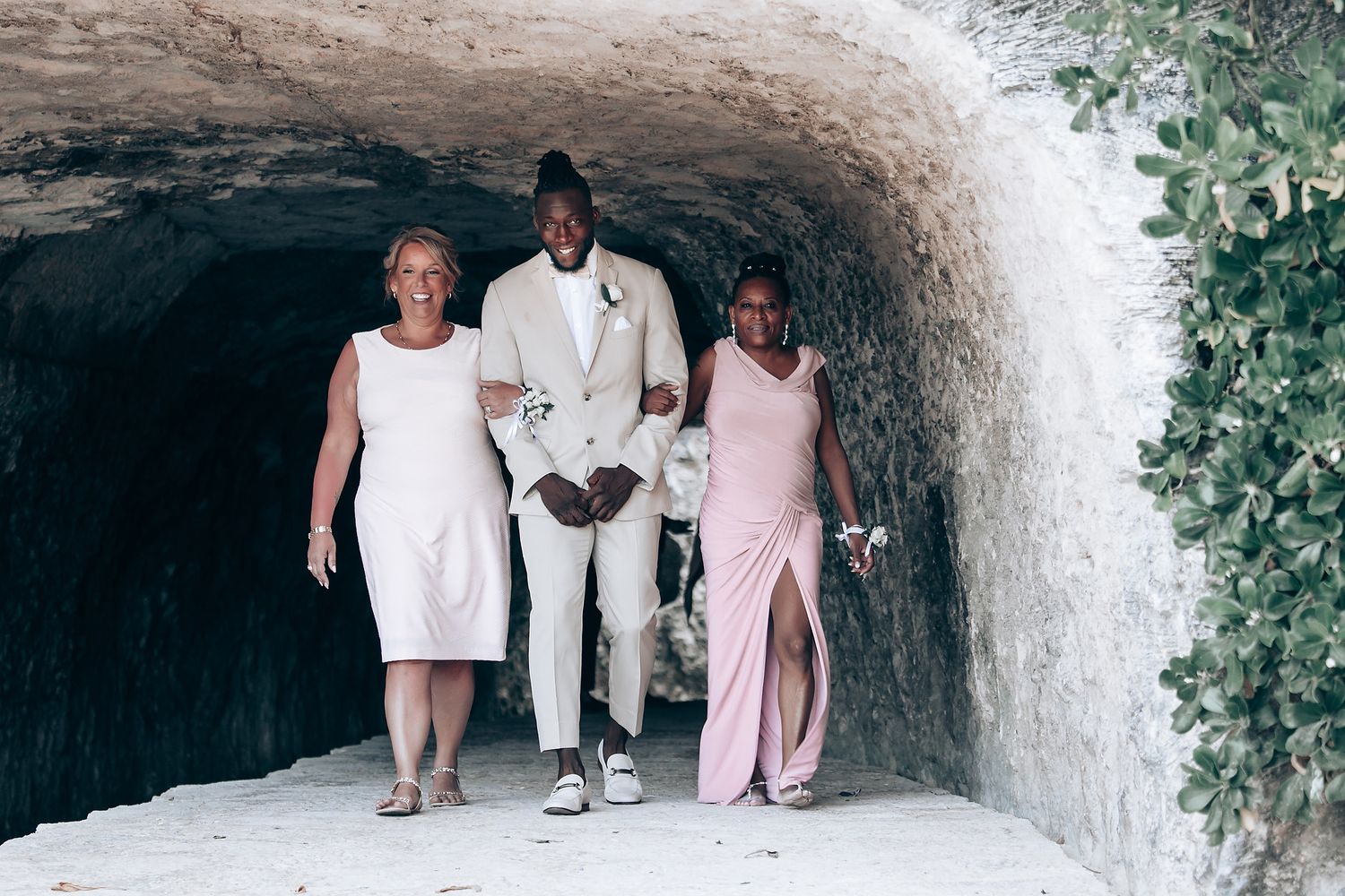 Wedding attendees walk through a stone archway tunnel decorated with greenery.