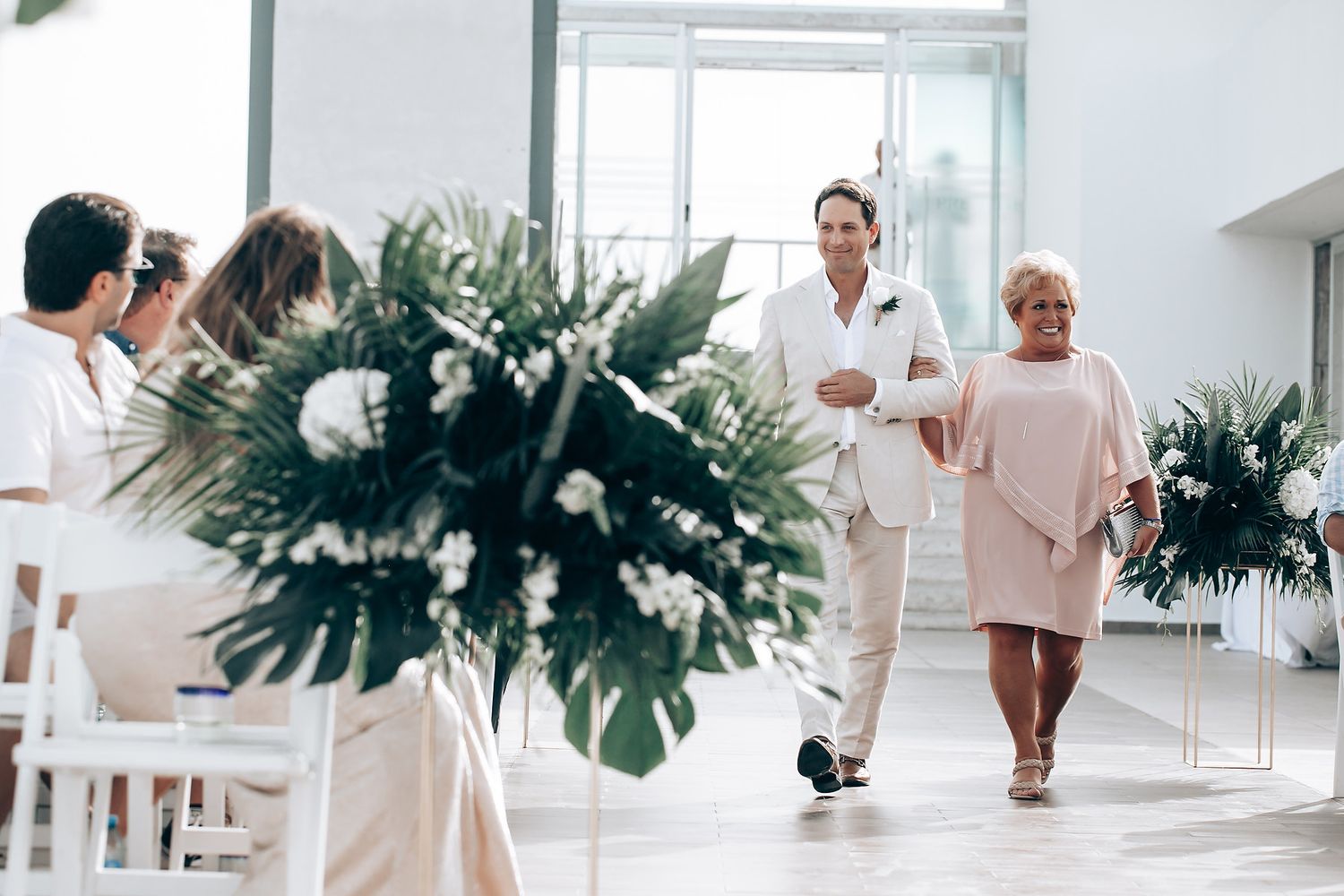 Modern wedding ceremony setup with tropical palm leaves and white chairs in a bright indoor space.