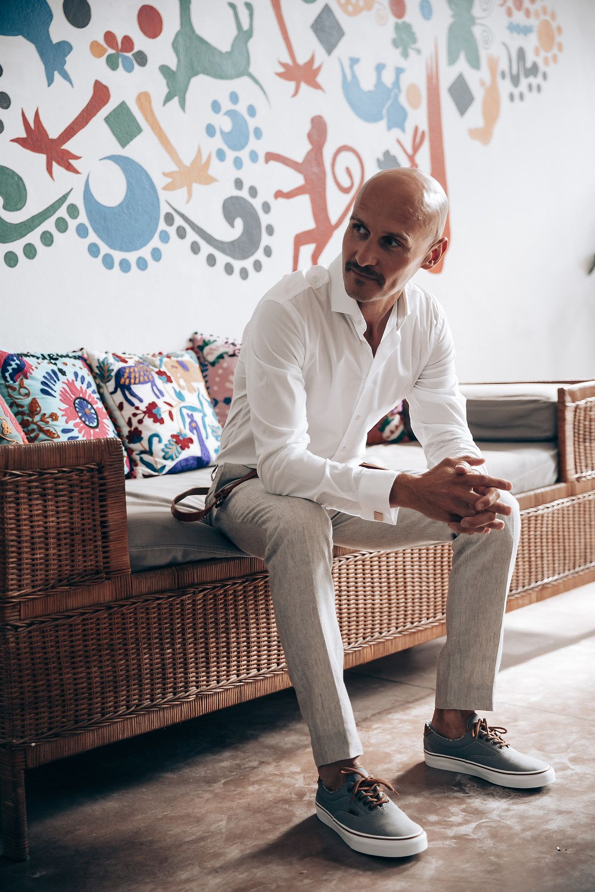 A person in white clothing sits on a wicker bench against a wall decorated with colorful artistic patterns.
