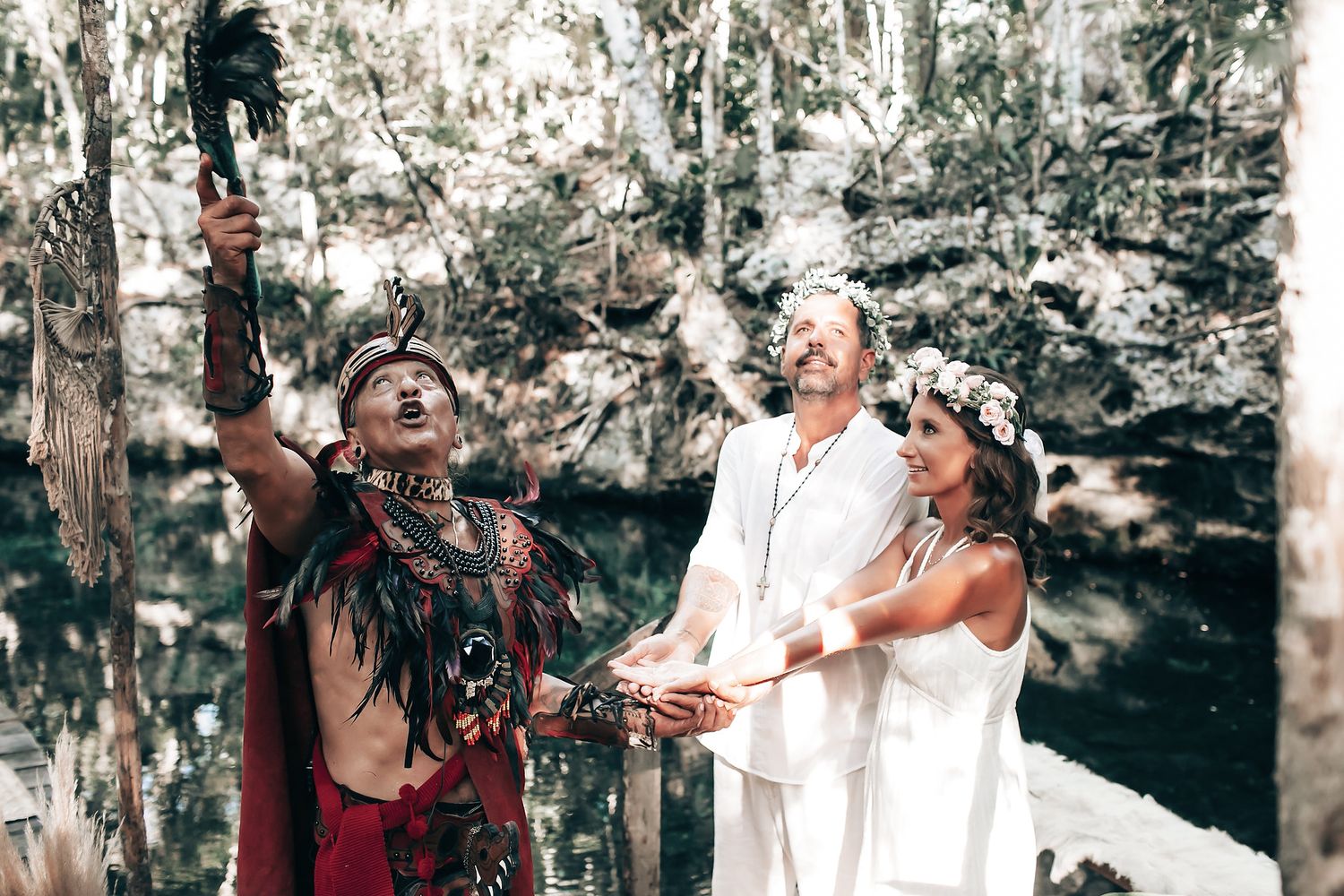 A couple stands in a lush, forested landscape, surrounded by greenery and a body of water.