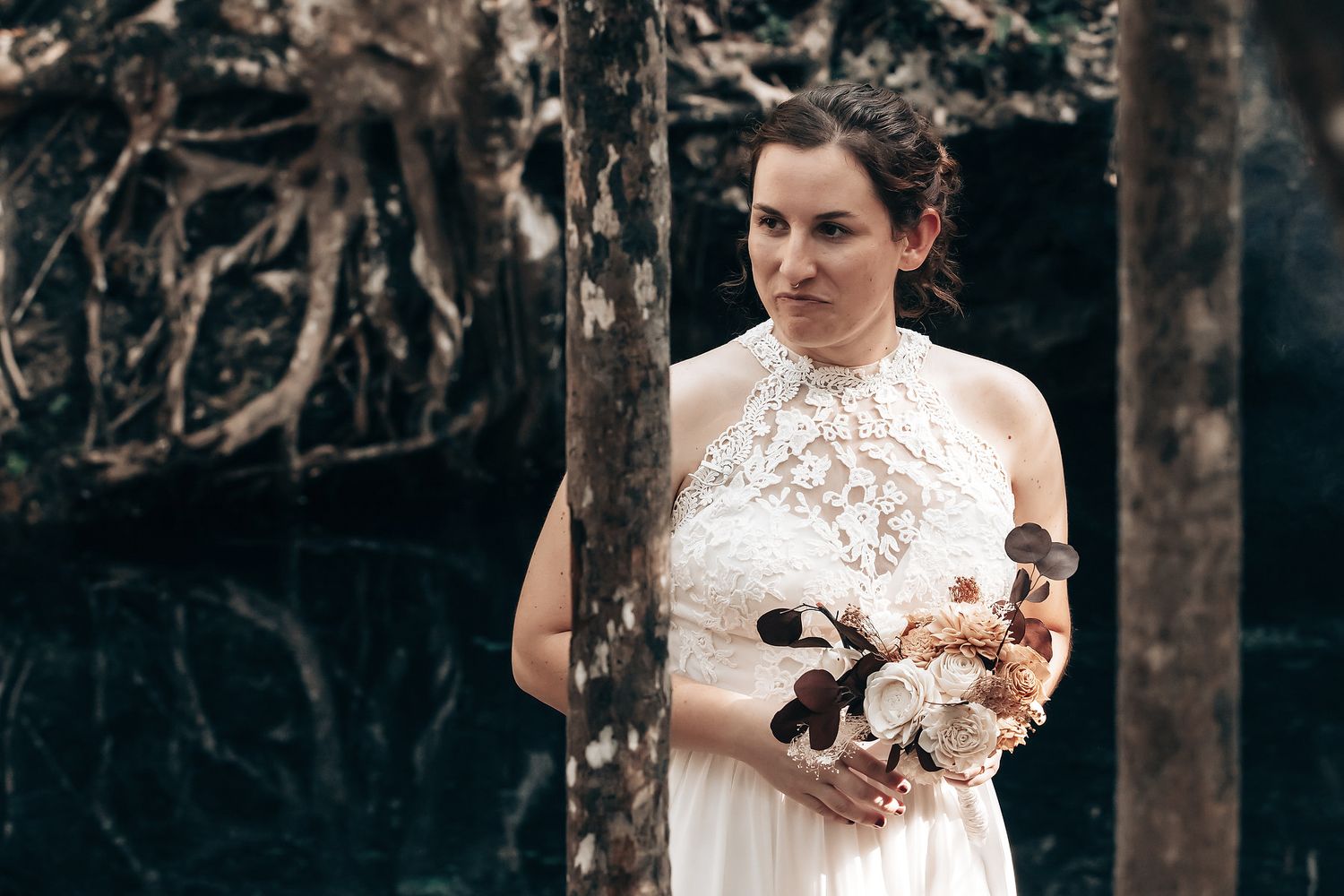 A woman in a white wedding dress stands among the trees in a forest setting.