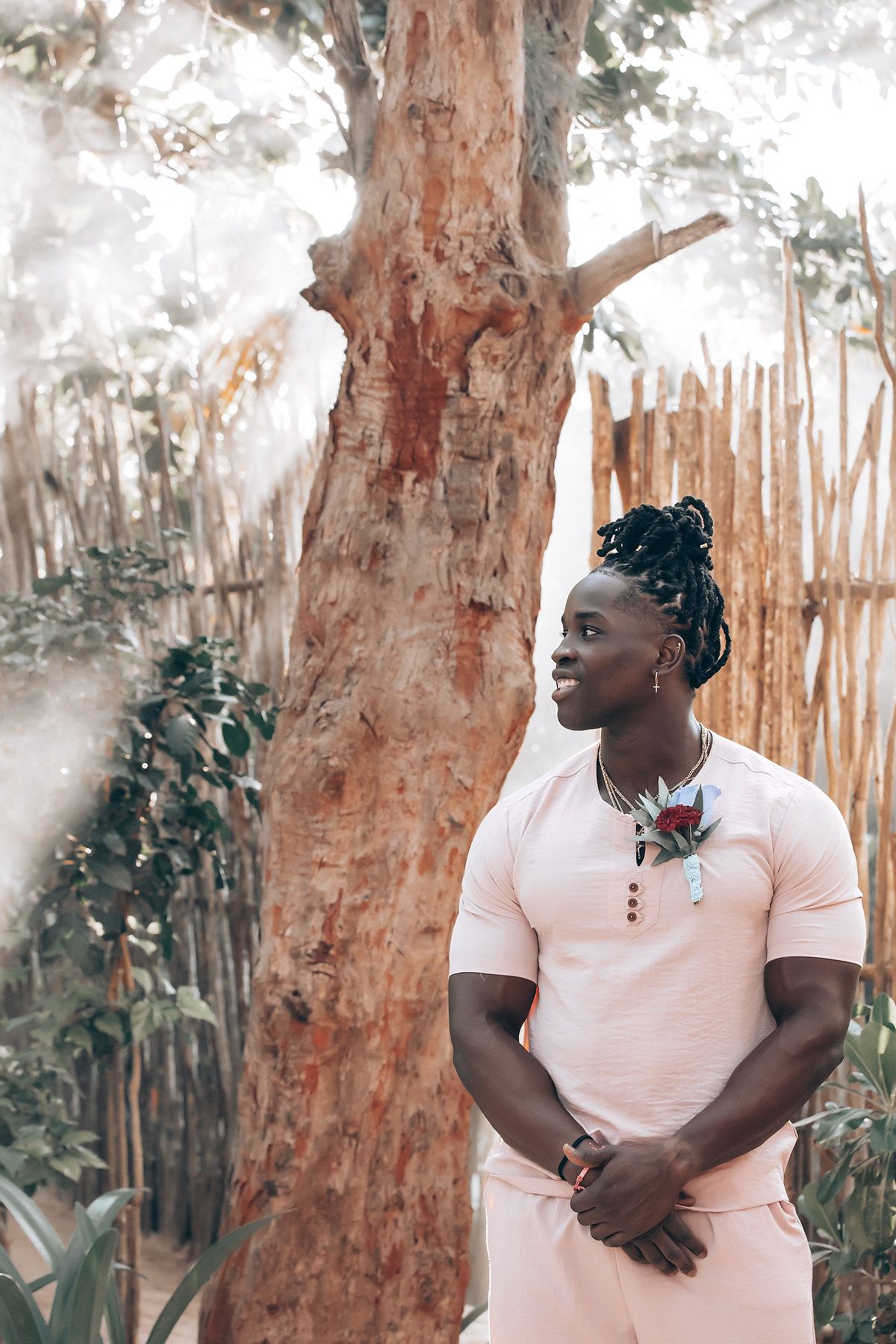 A man stands in a lush outdoor setting, wearing a pink shirt.