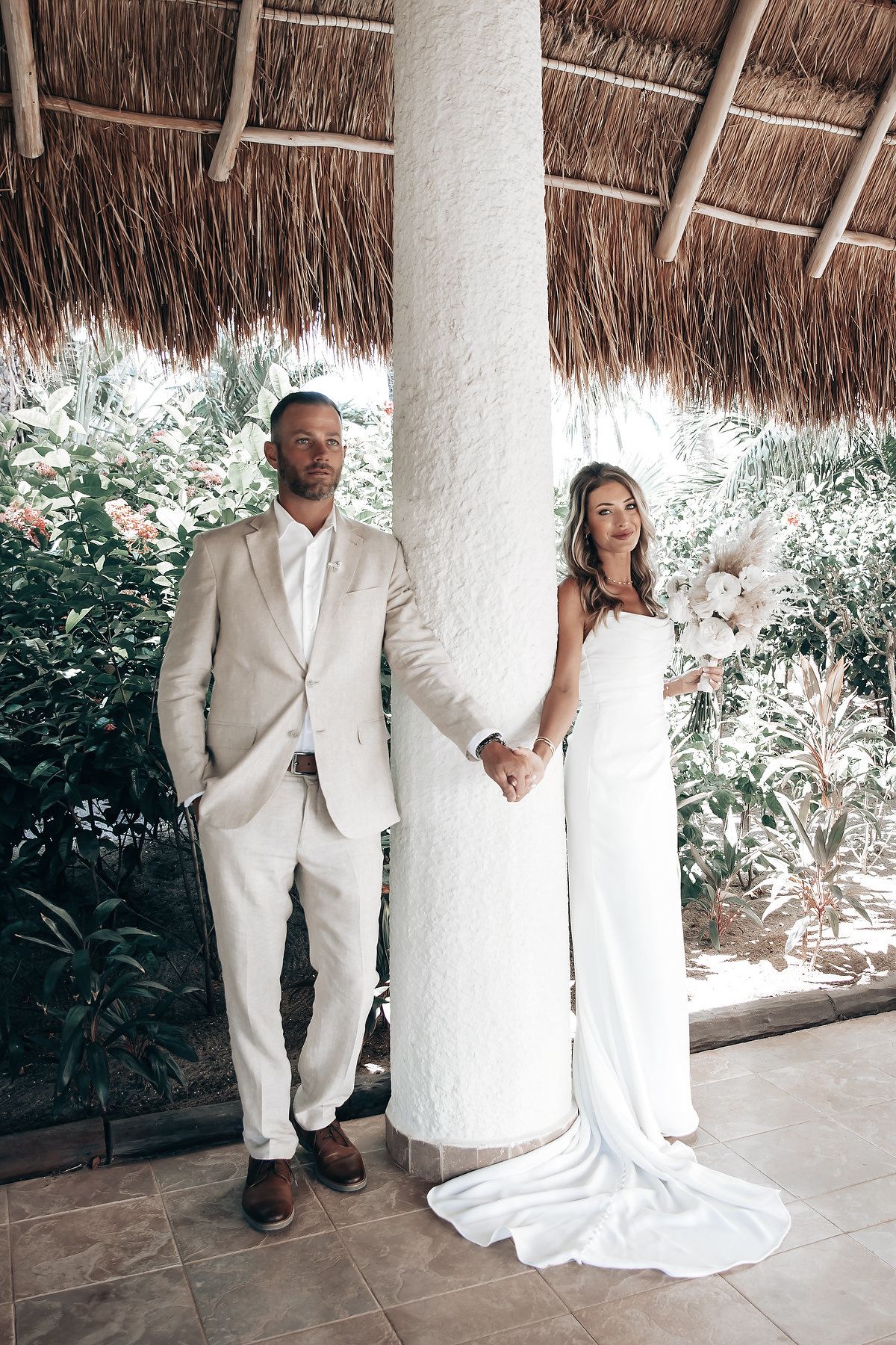 A bride and groom stand together in a tropical garden setting, holding hands and smiling.