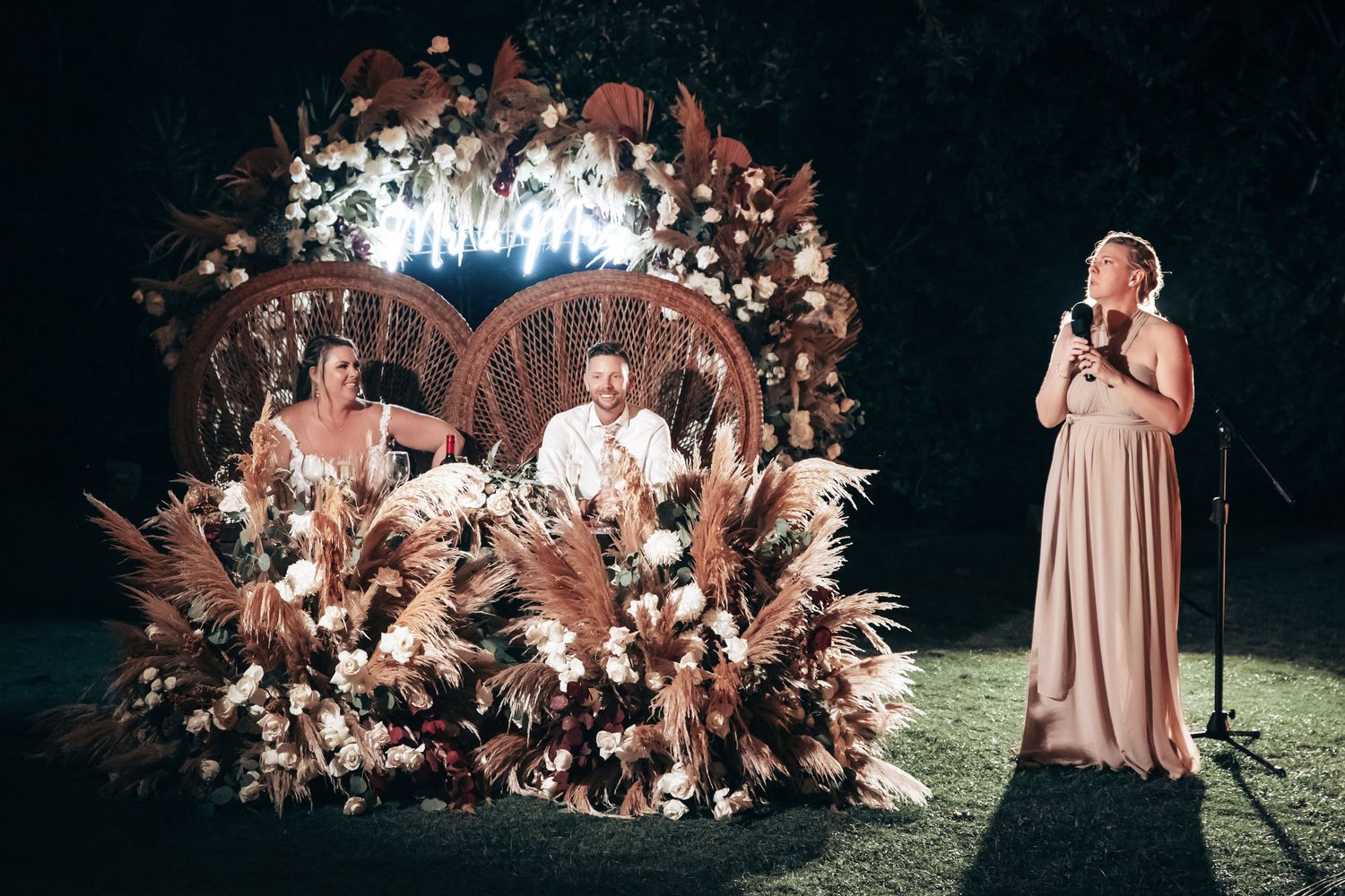 A singer performs at a romantic outdoor wedding reception as the newlyweds sit behind a lush floral arrangement.