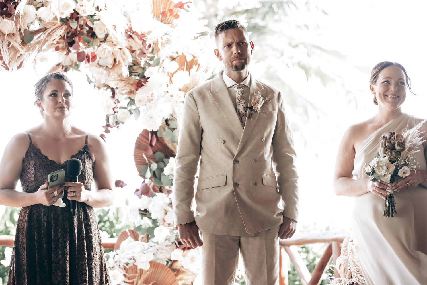 Groom in beige suit standing at wedding altar flanked by bridesmaids with bouquets.