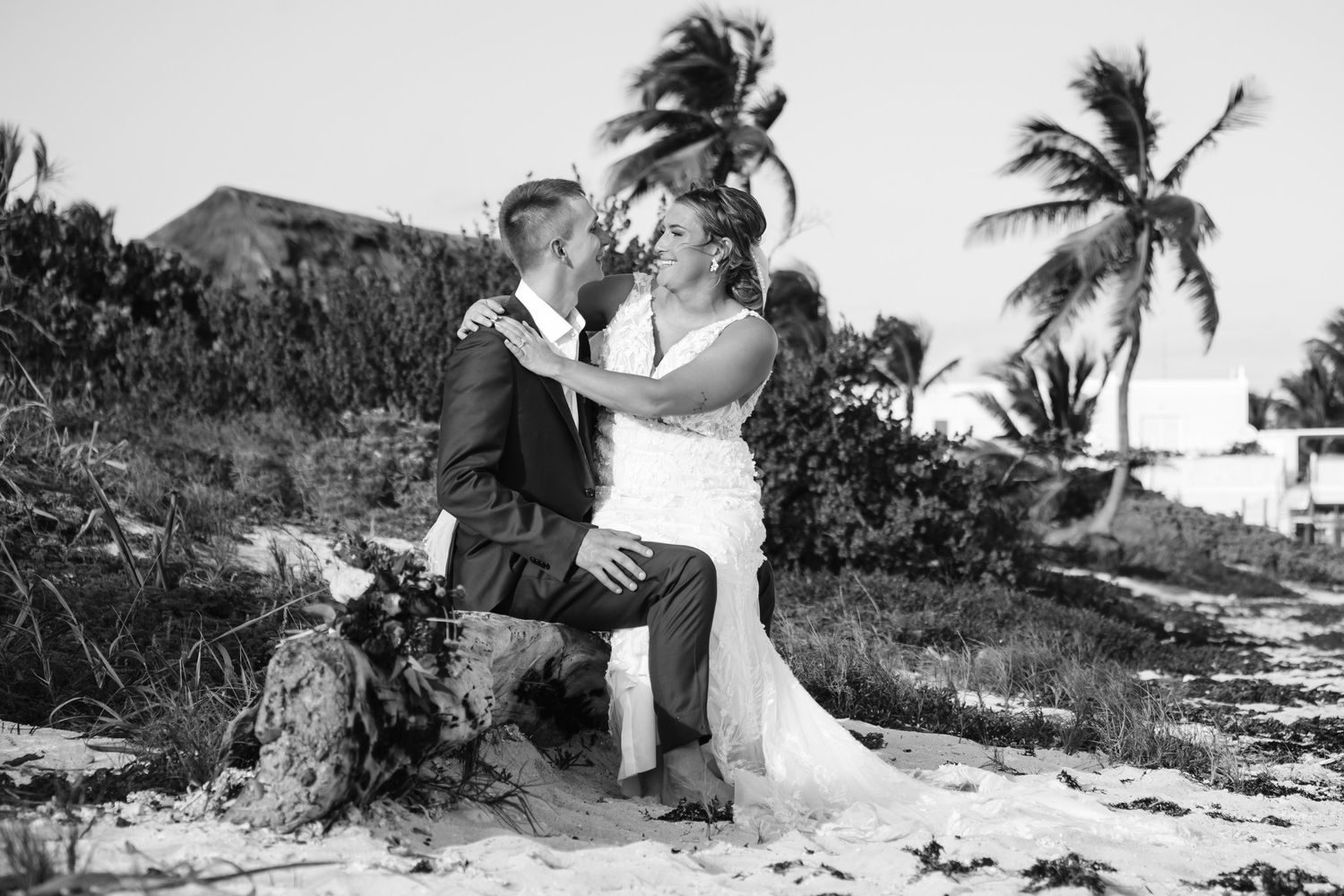 Black and white photo of bride and groom sharing a kiss near palm trees on sandy beach.