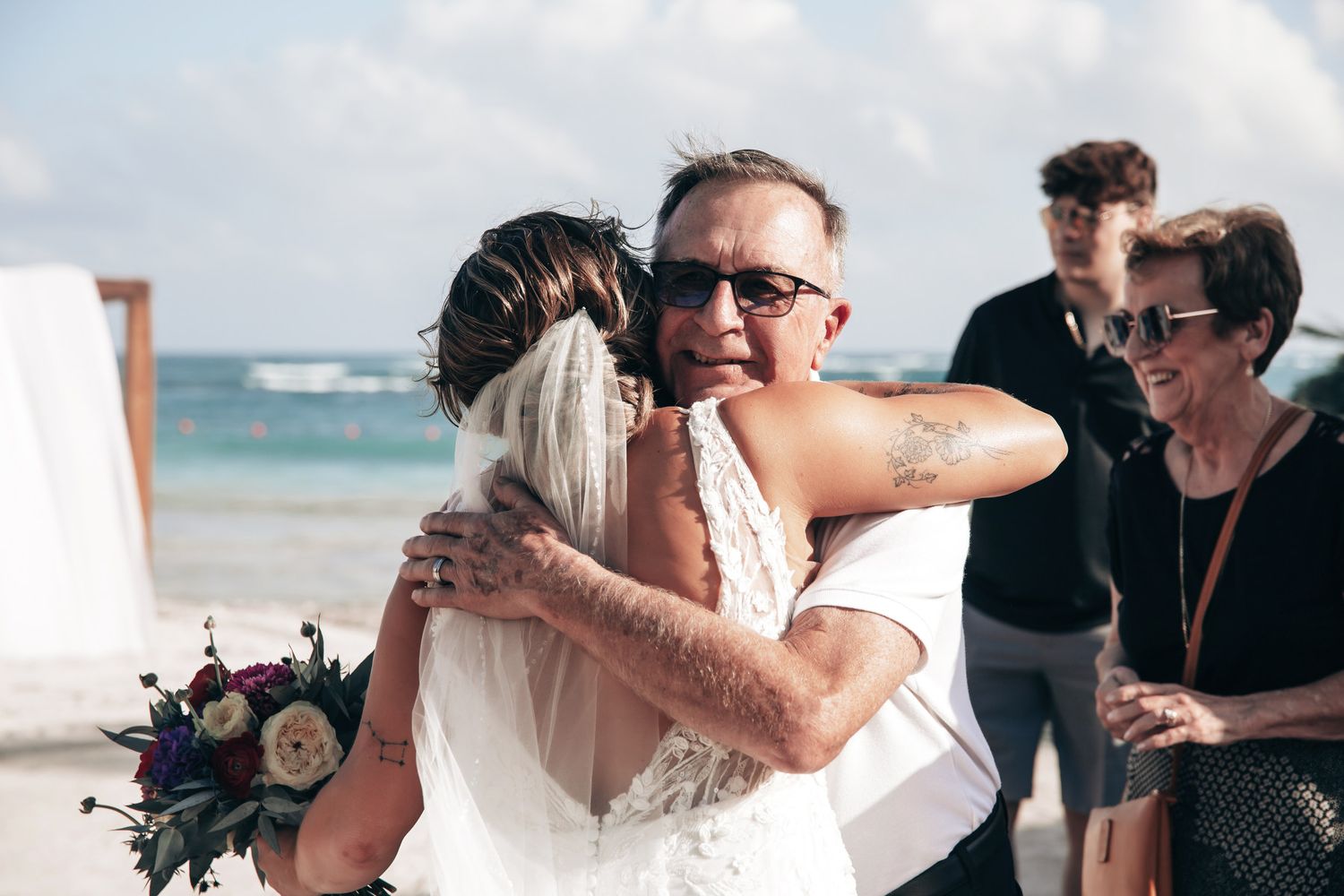Bride in white lace gown embraces emotional father on beach wedding venue with ocean backdrop.